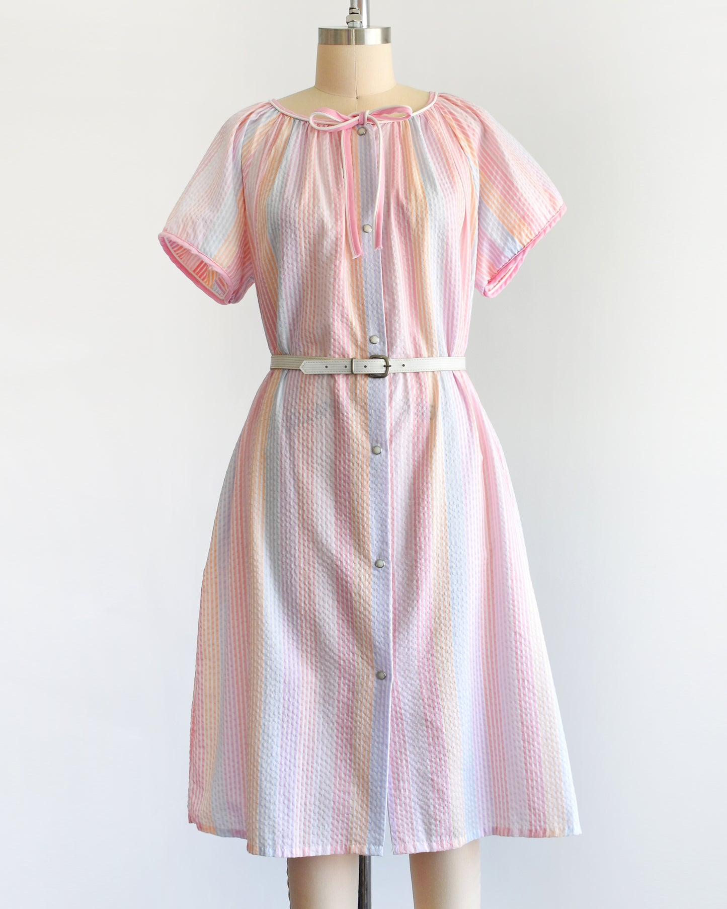 a vintage 1980s house dress that  features pastel rainbow stripes in blue, pink, orange, purple and white, along with a pink and white bow tie at the neck, and pearl snap buttons down the front. There is a belt around the waist in this photo