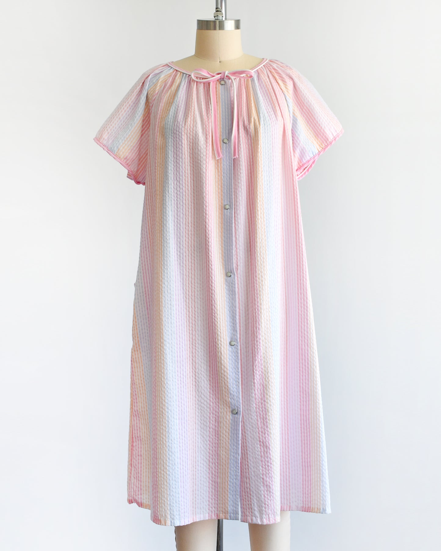 a vintage 1980s house dress that  features pastel rainbow stripes in blue, pink, orange, purple and white, along with a pink and white bow tie at the neck, and pearl snap buttons down the front. 