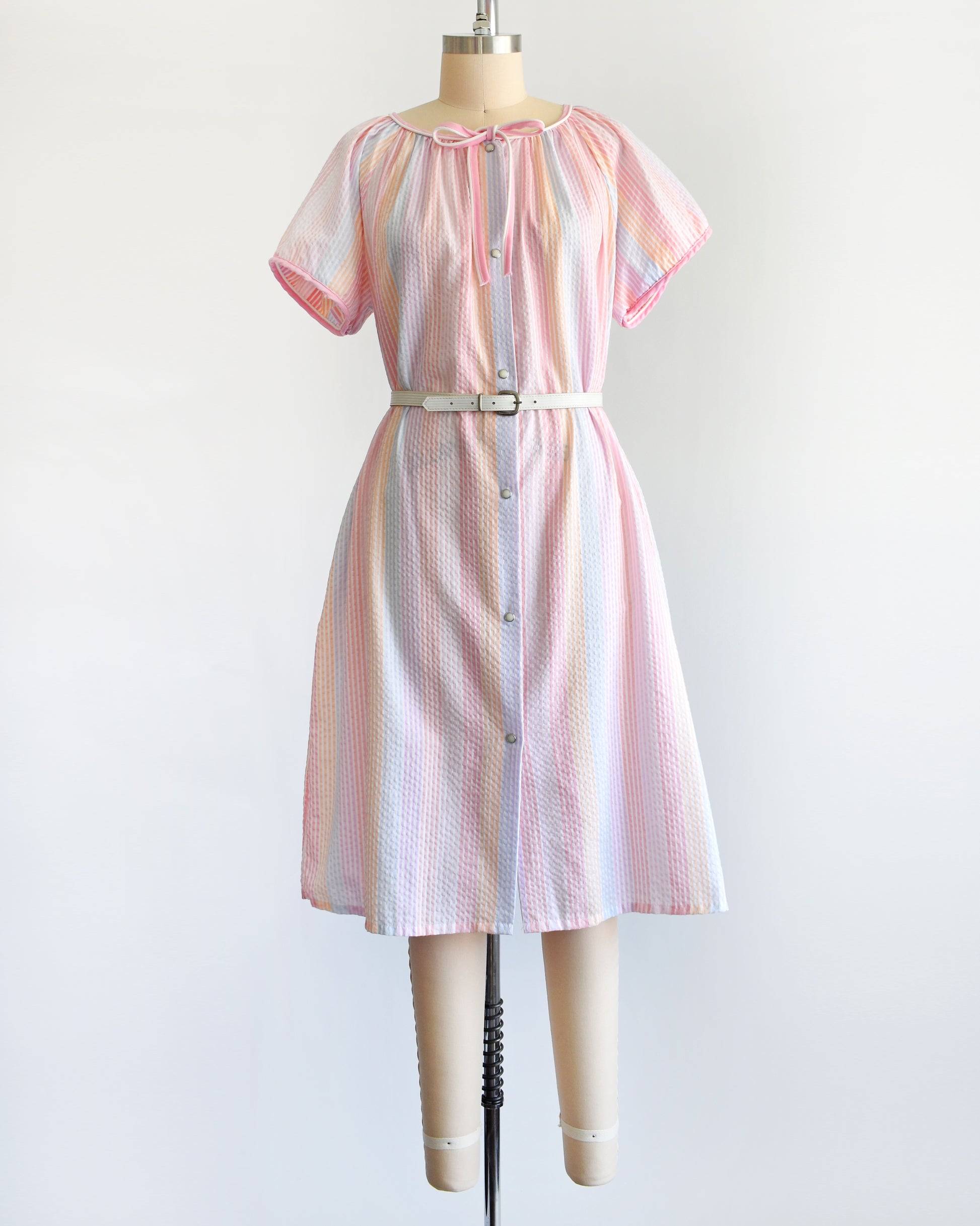 a vintage 1980s house dress that  features pastel rainbow stripes in blue, pink, orange, purple and white, along with a pink and white bow tie at the neck, and pearl snap buttons down the front. There is a belt around the waist in the photo