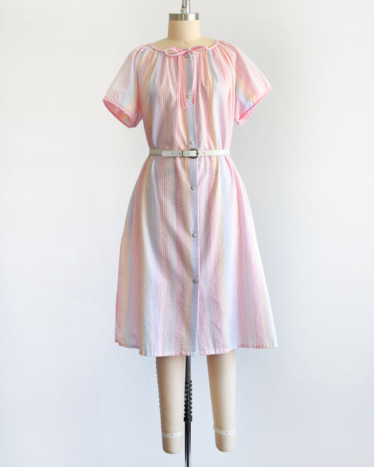 a vintage 1980s house dress that  features pastel rainbow stripes in blue, pink, orange, purple and white, along with a pink and white bow tie at the neck, and pearl snap buttons down the front. There is a belt around the waist in the photo