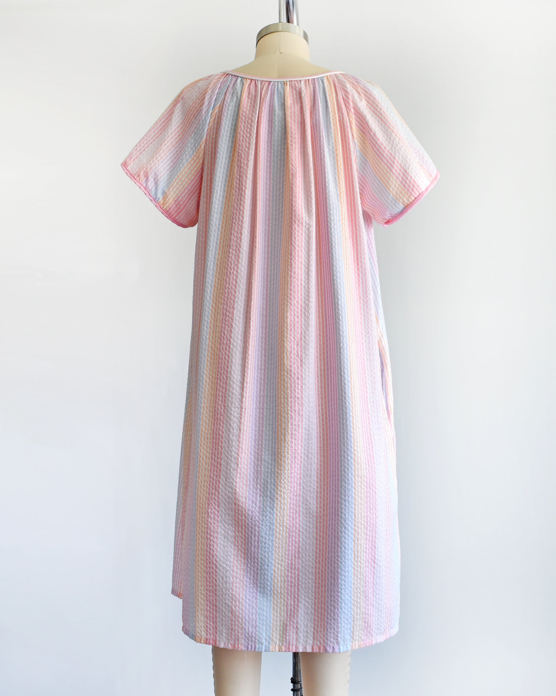 back view of a vintage 1980s house dress that  features pastel rainbow stripes in blue, pink, orange, purple and white, along with a pink and white bow tie at the neck, and pearl snap buttons down the front. 