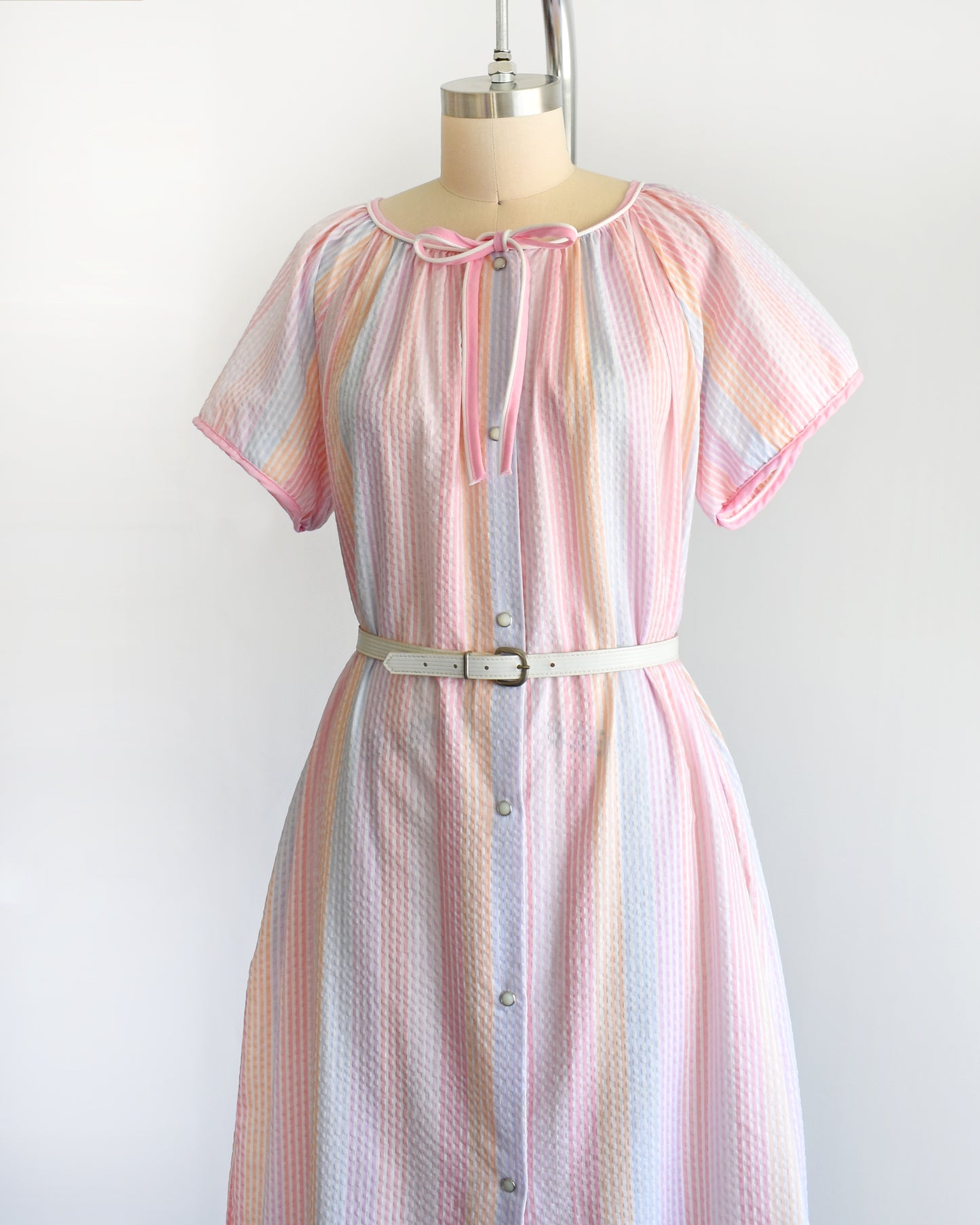 side front view of a vintage 1980s house dress that  features pastel rainbow stripes in blue, pink, orange, purple and white, along with a pink and white bow tie at the neck, and pearl snap buttons down the front.  there is a belt around the waist