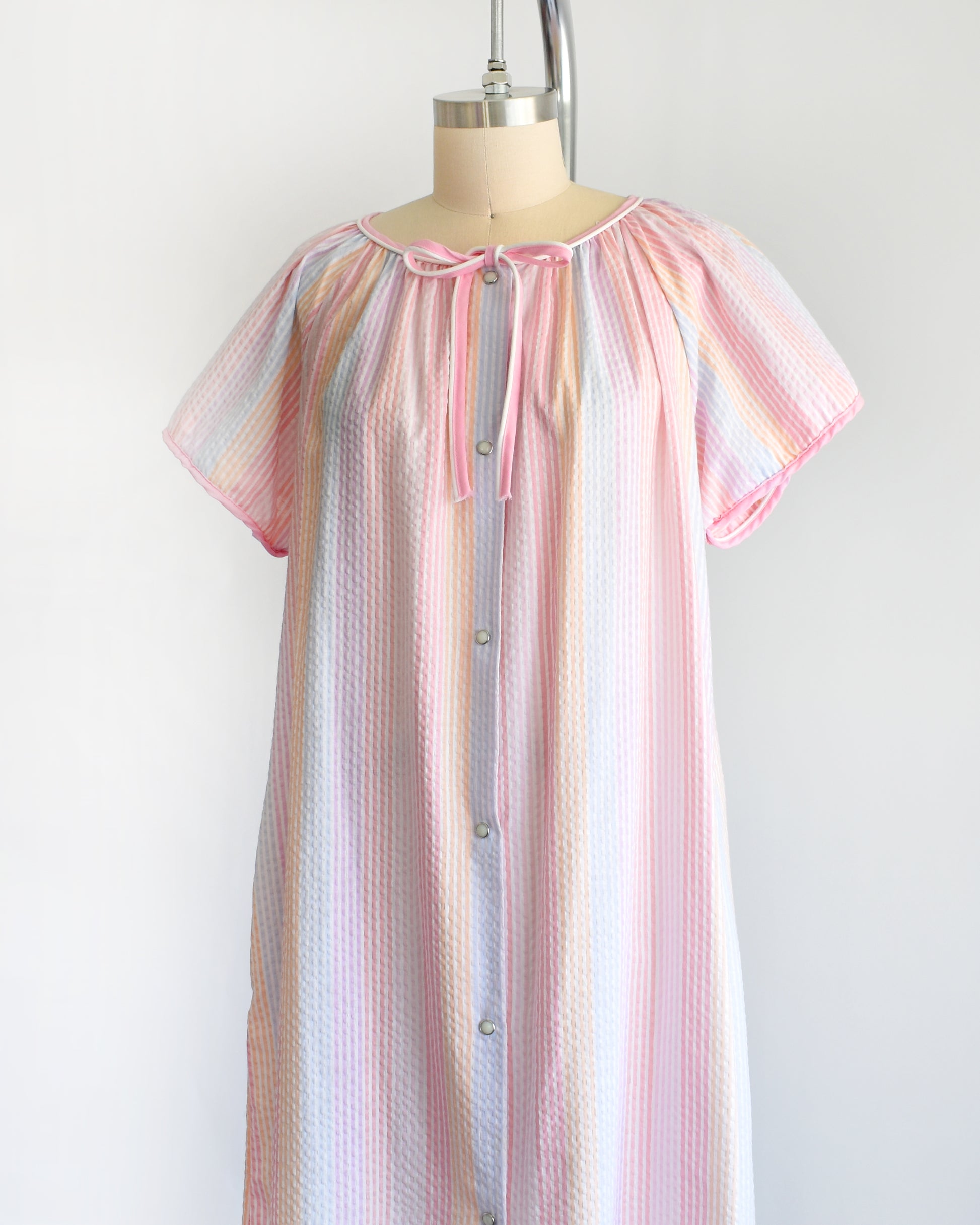 side front view of a vintage 1980s house dress that  features pastel rainbow stripes in blue, pink, orange, purple and white, along with a pink and white bow tie at the neck, and pearl snap buttons down the front. 