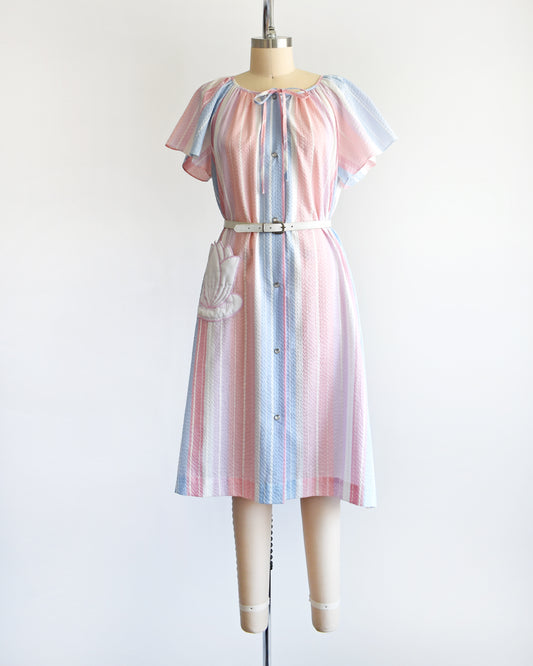 A vintage 1980s blue, pink, and white striped house dress with a white lily pad pocket on a dress form. There is a white belt around the waist which is not included.