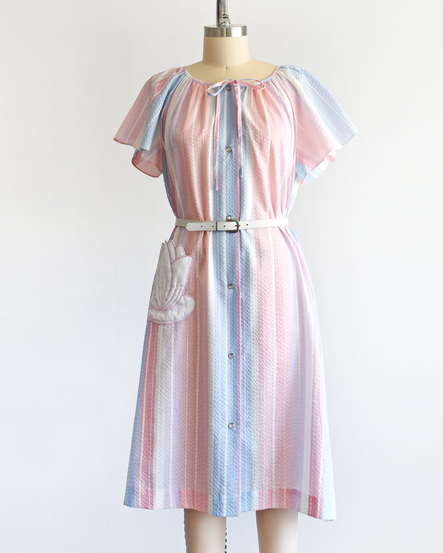 A vintage 1980s blue, pink, and white striped house dress with a white lily pad pocket on a dress form. There is a white belt around the waist which is not included.