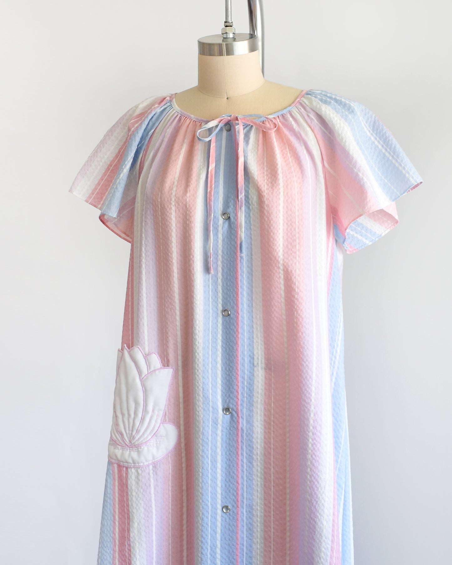 side front view of a vintage 1980s blue, pink, and white striped house dress with a white lily pad pocket on a dress form