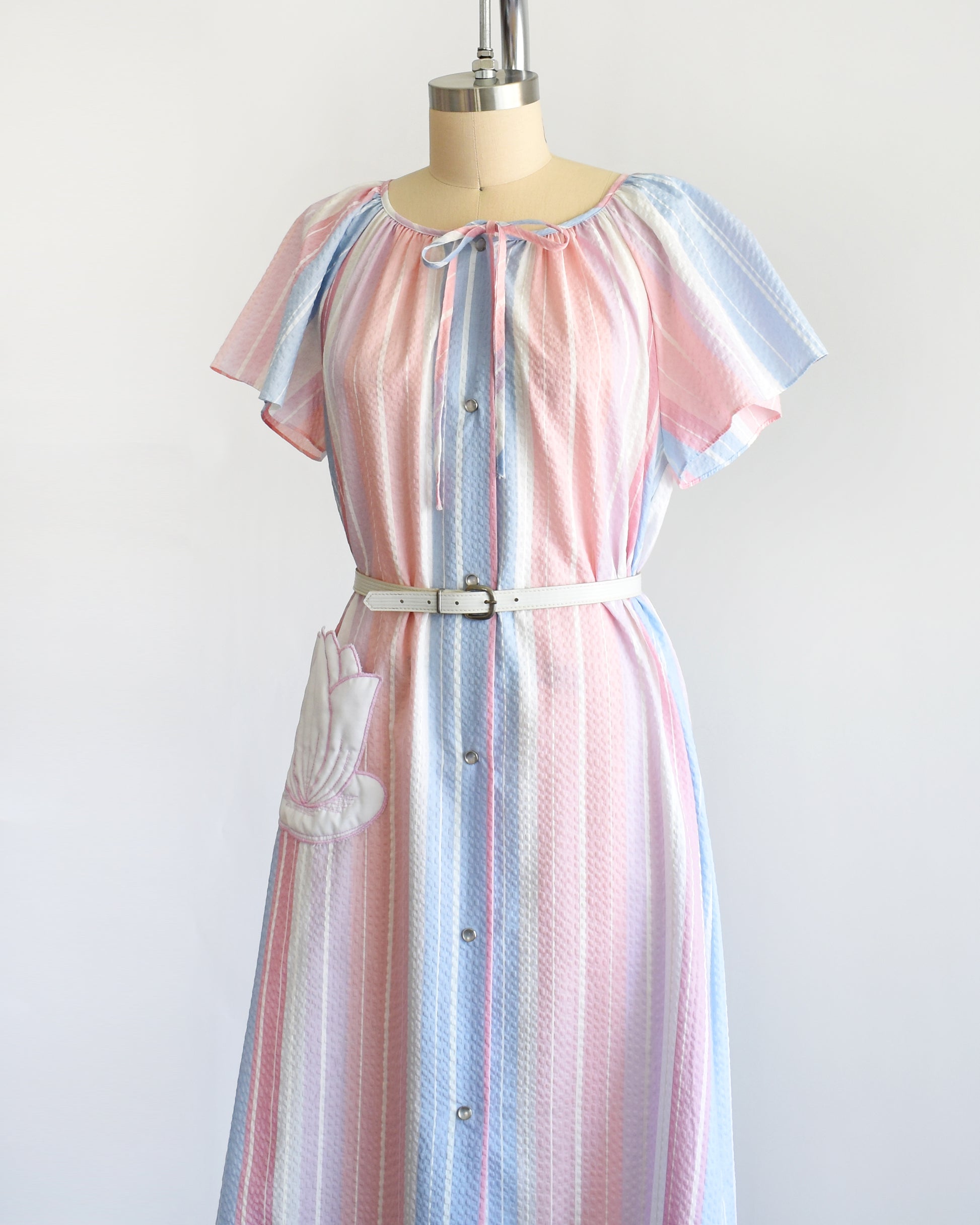 side front view of a vintage 1980s blue, pink, and white striped house dress with a white lily pad pocket on a dress form. There is a white belt around the waist which is not included.