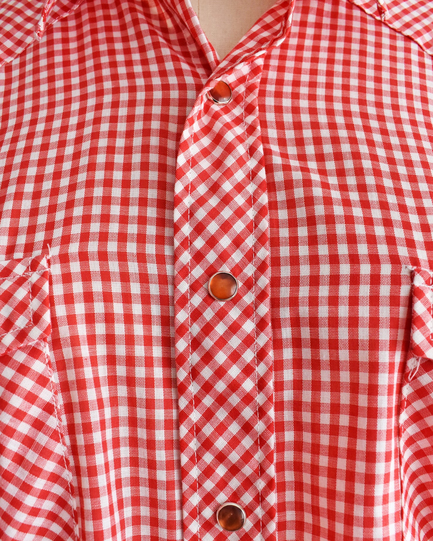 close up of the red pearl snaps on a vintage 1970s red and white gingham shirt on a mannequin against a white background