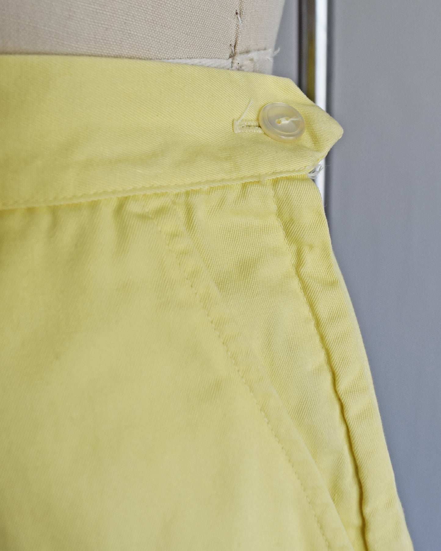 close up of the button on vintage 1960s yellow high-waisted shorts displayed on a mannequin against a white  background.