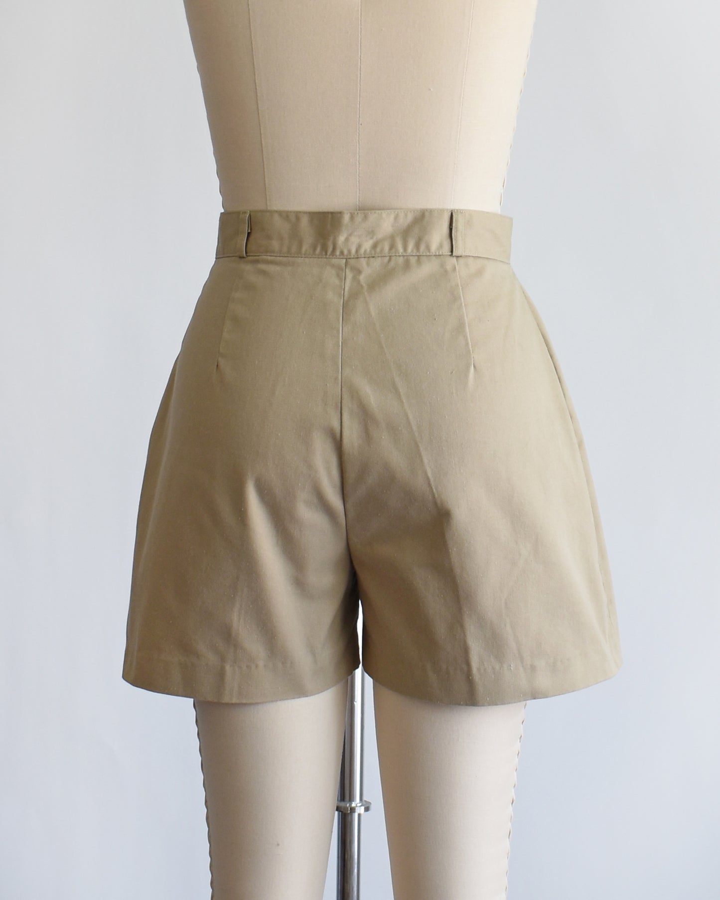 back view of a pair of beige high-waisted shorts displayed on a cream dress form mannequin against a plain white background.