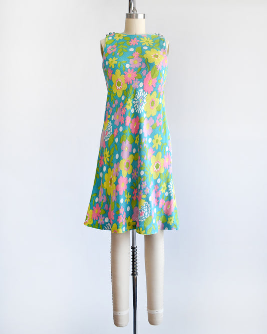 A vintage 1960s blue dress that has a flower power print in pinks, yellows, greens, and white. The dress is on a dress form.