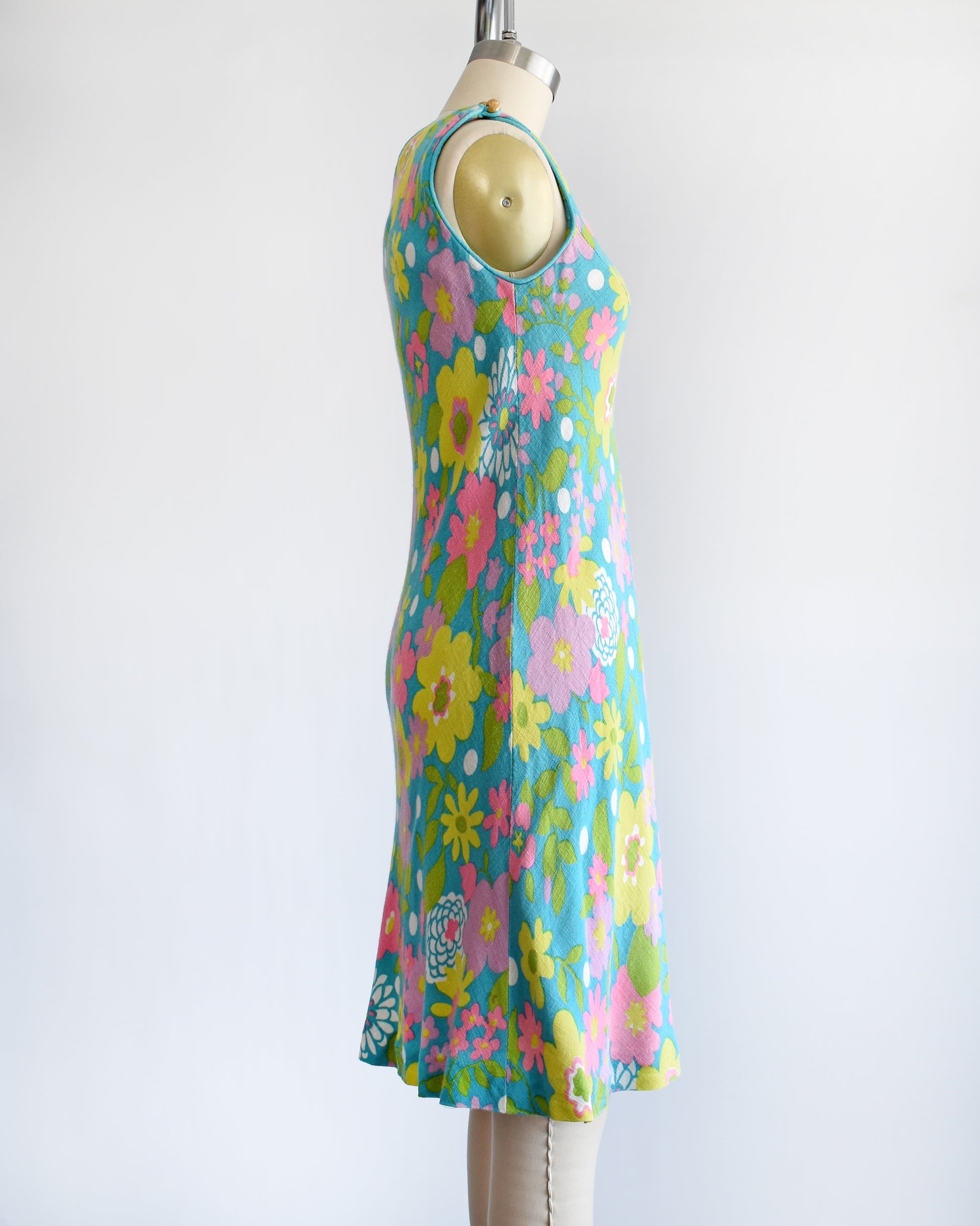 Side view of a vintage 1960s blue dress that has a flower power print in pinks, yellows, greens, and white. The dress is on a dress form.