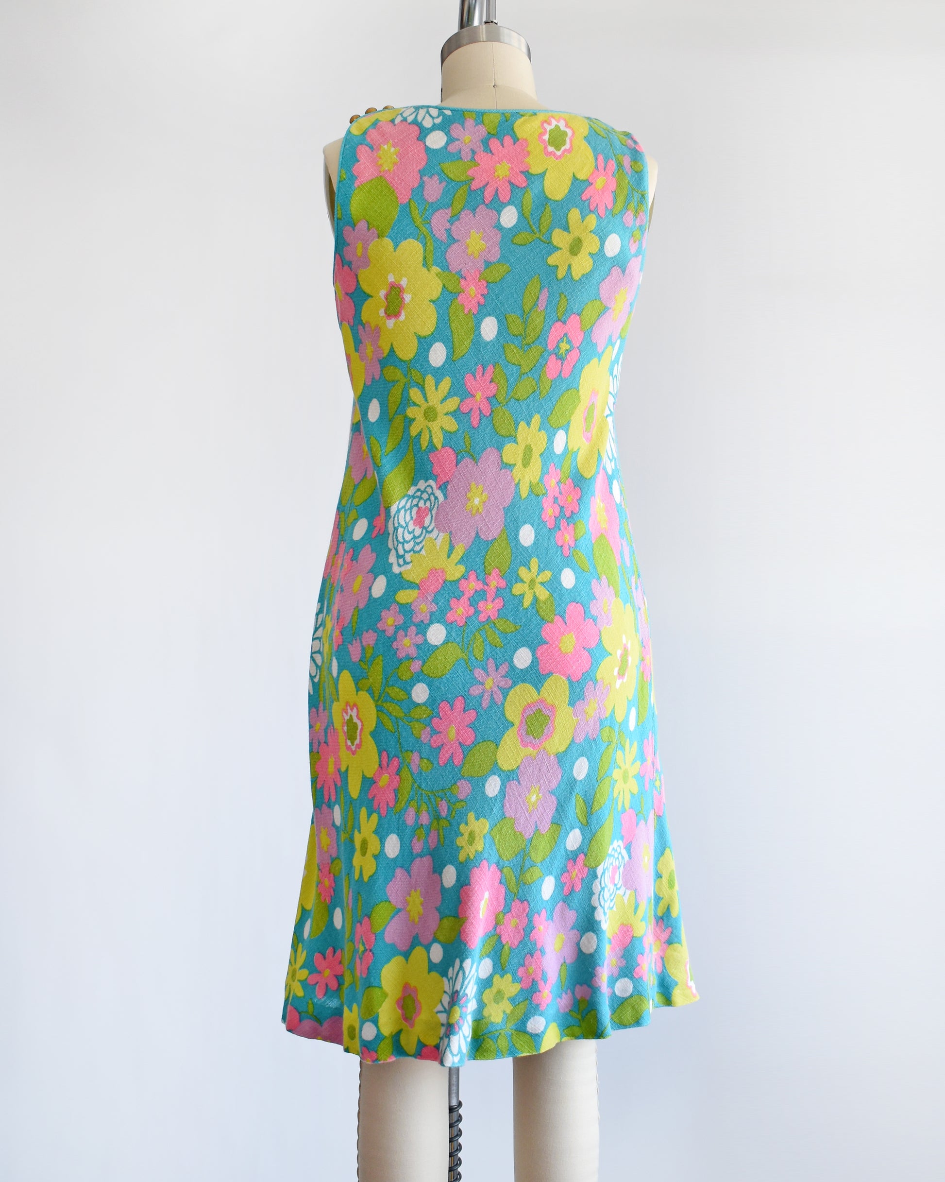 Back view of a vintage 1960s blue dress that has a flower power print in pinks, yellows, greens, and white. The dress is on a dress form.