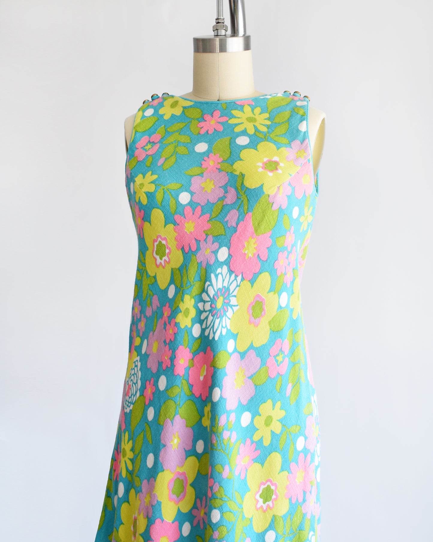 Side front view of a vintage 1960s blue dress that has a flower power print in pinks, yellows, greens, and white. The dress is on a dress form.