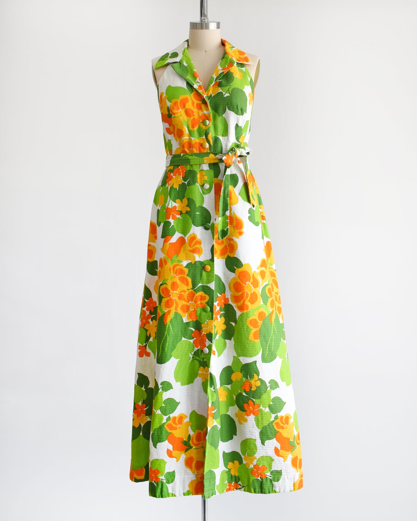 a vintage 1970s white maxi dress by Malia that has a large orange and green floral print, collared neckline, button front, and matching belt.