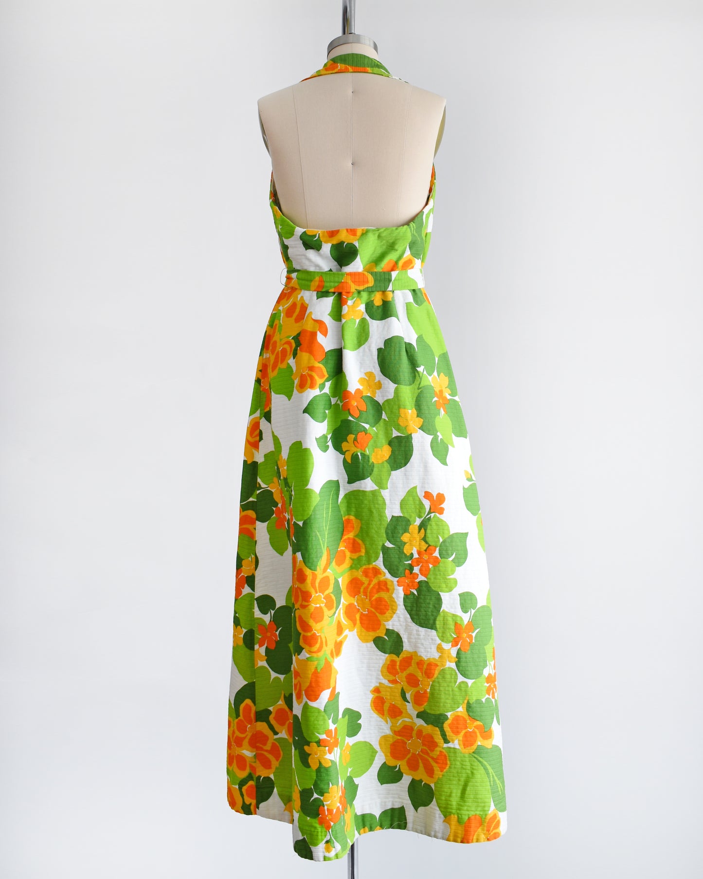 back view of a vintage 1970s white maxi dress by Malia that has a large orange and green floral print, collared neckline, button front, and matching belt.