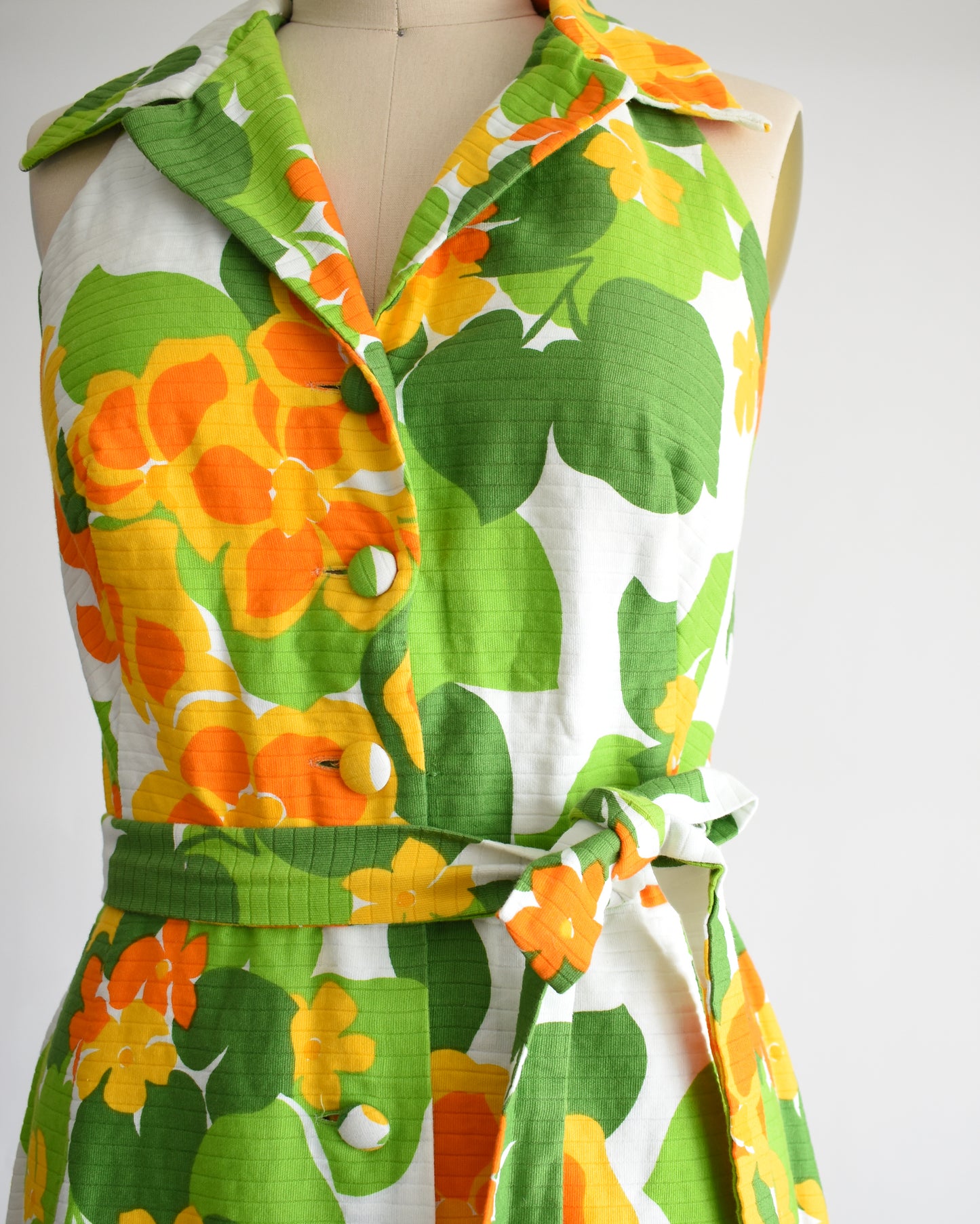 close up of the bodice of a vintage 1970s white maxi dress by Malia that has a large orange and green floral print, collared neckline, button front, and matching belt.