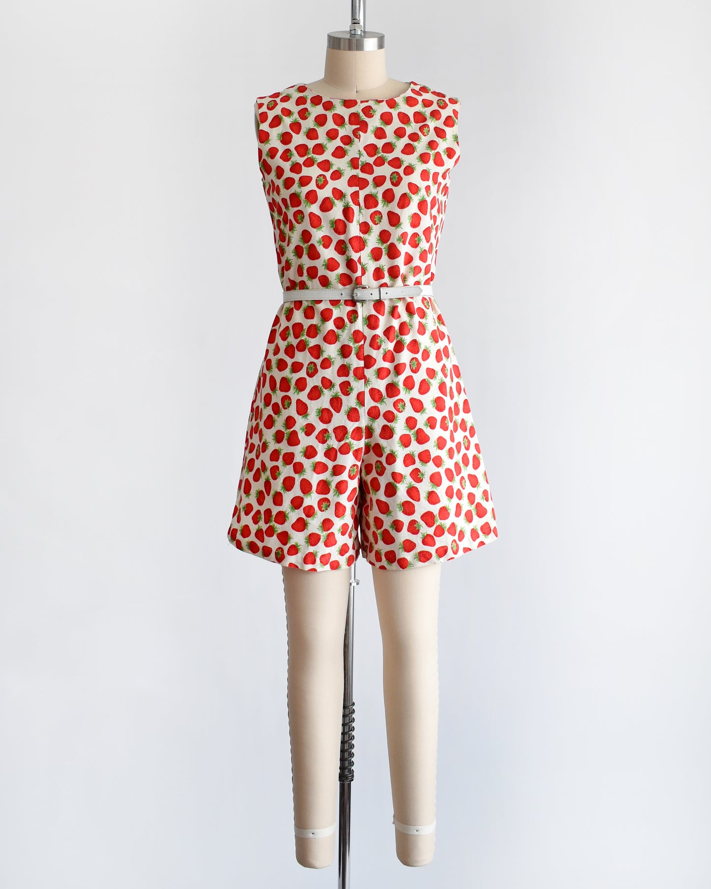 a vintage 1960s white romper with a red and green strawberry print. the romper is modeled with a white belt (not included) and is on a dress form.