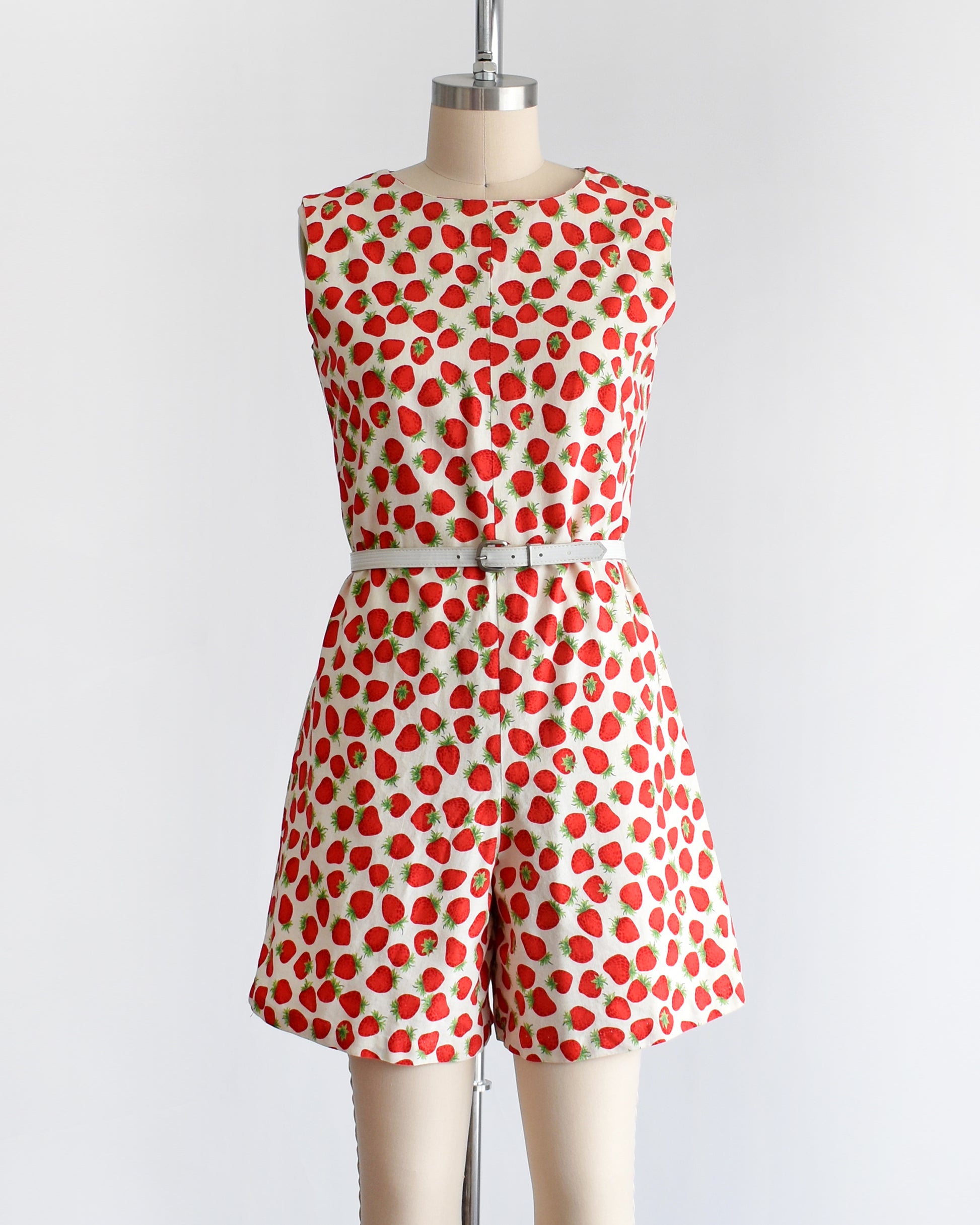 a vintage 1960s white romper with a red and green strawberry print. the romper is modeled with a white belt (not included) and is on a dress form.