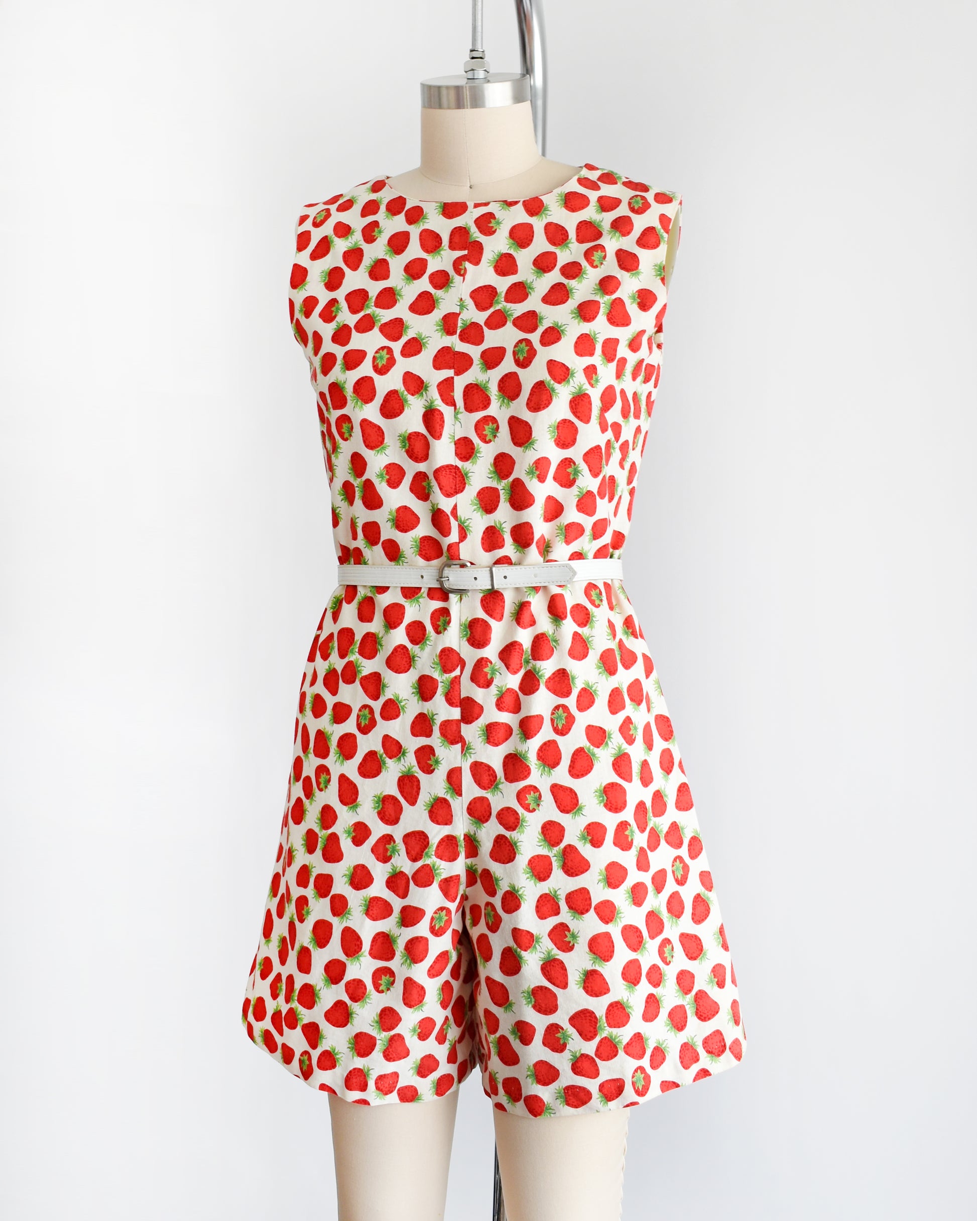 side front view of a vintage 1960s white romper with a red and green strawberry print. the romper is modeled with a white belt (not included) and is on a dress form.