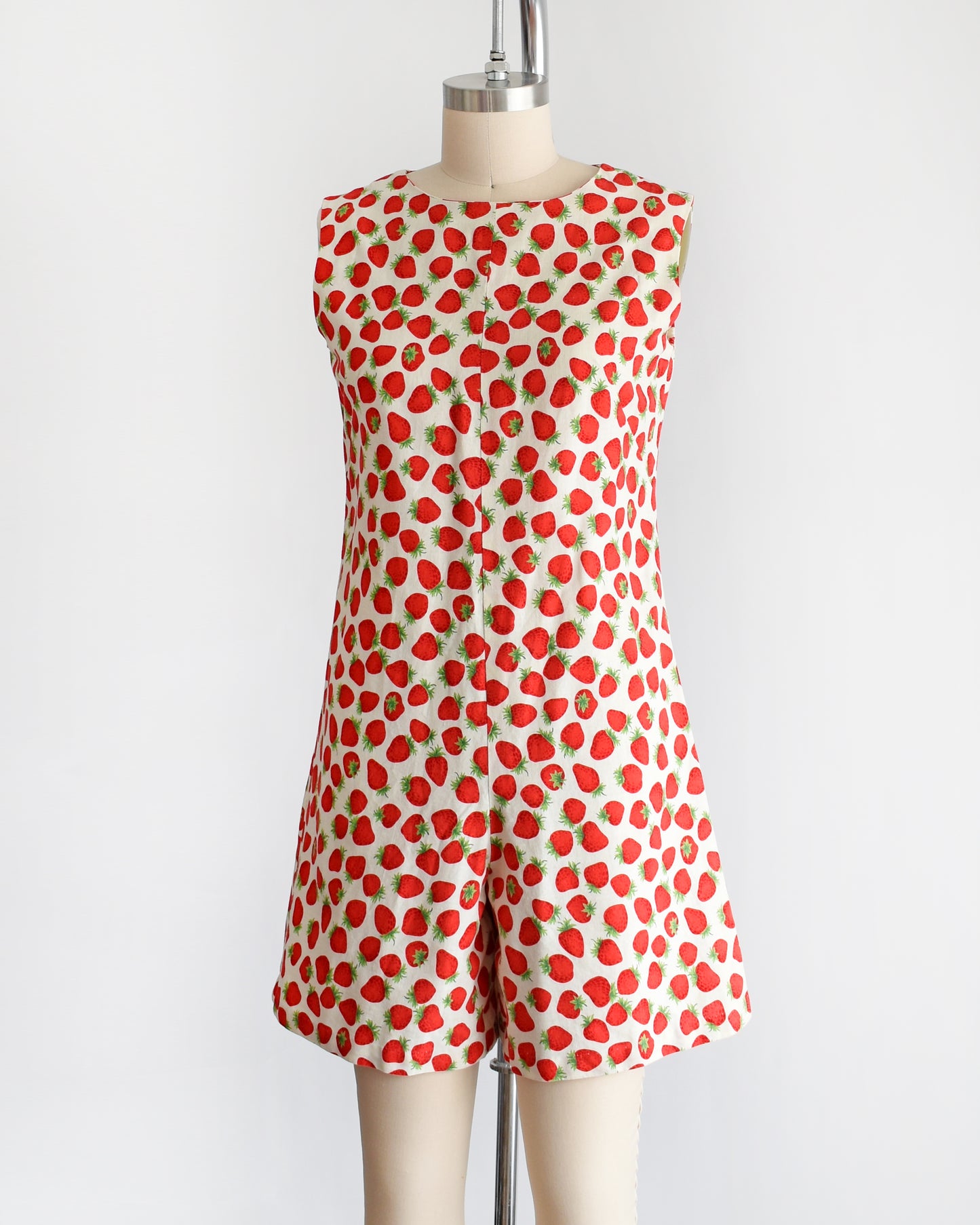 a vintage 1960s white romper with a red and green strawberry print. the romper is modeled on a dress form.