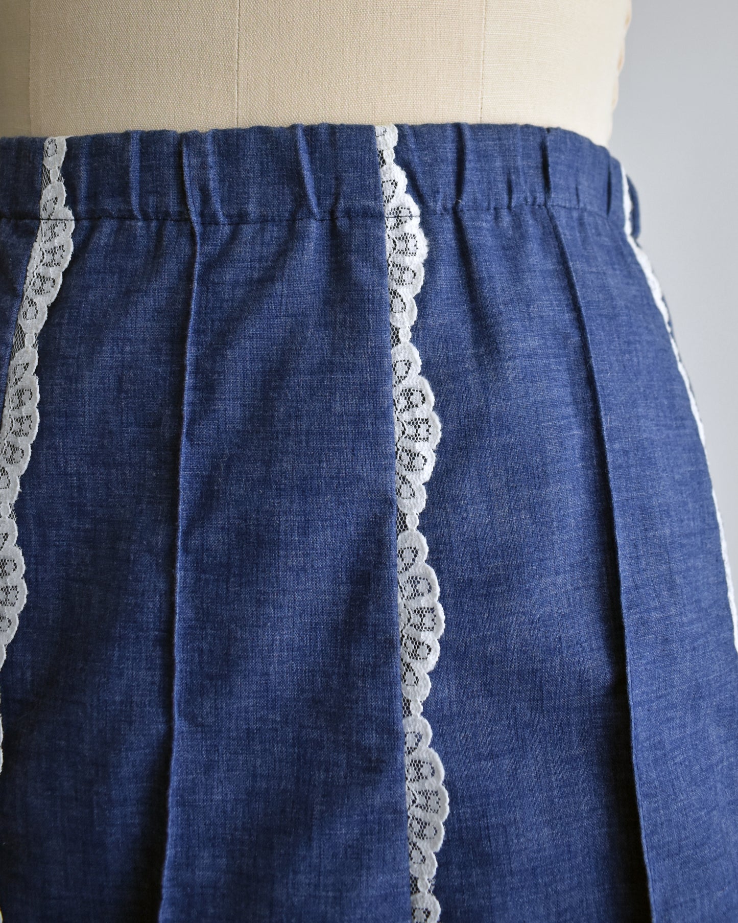 close up of the elastic waist of a vintage 1970s blue chambray lace skirt on a dress form against a white background