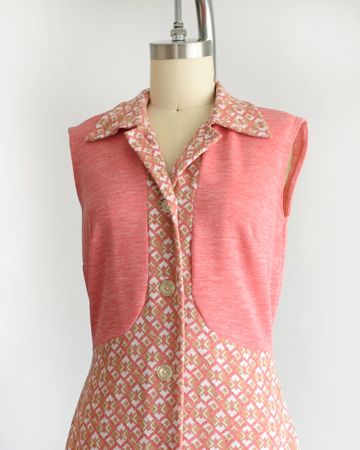 side front view of a vintage 1960s/1970s pink sleeveless dress with a geometric pattern on a mannequin against a white background. the top button is unbuttoned in this photo