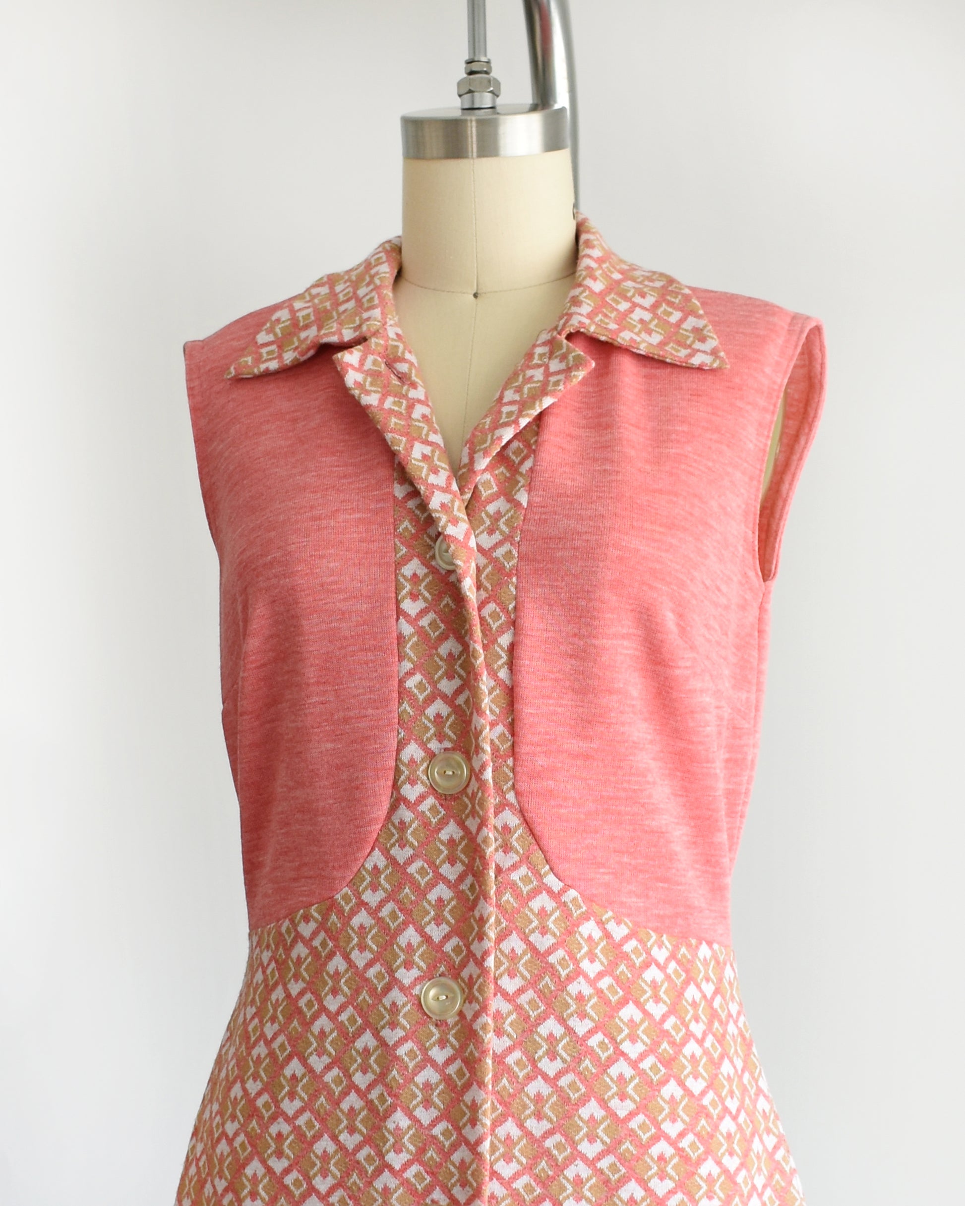 side front view of a vintage 1960s/1970s pink sleeveless dress with a geometric pattern on a mannequin against a white background. the top button is unbuttoned in this photo