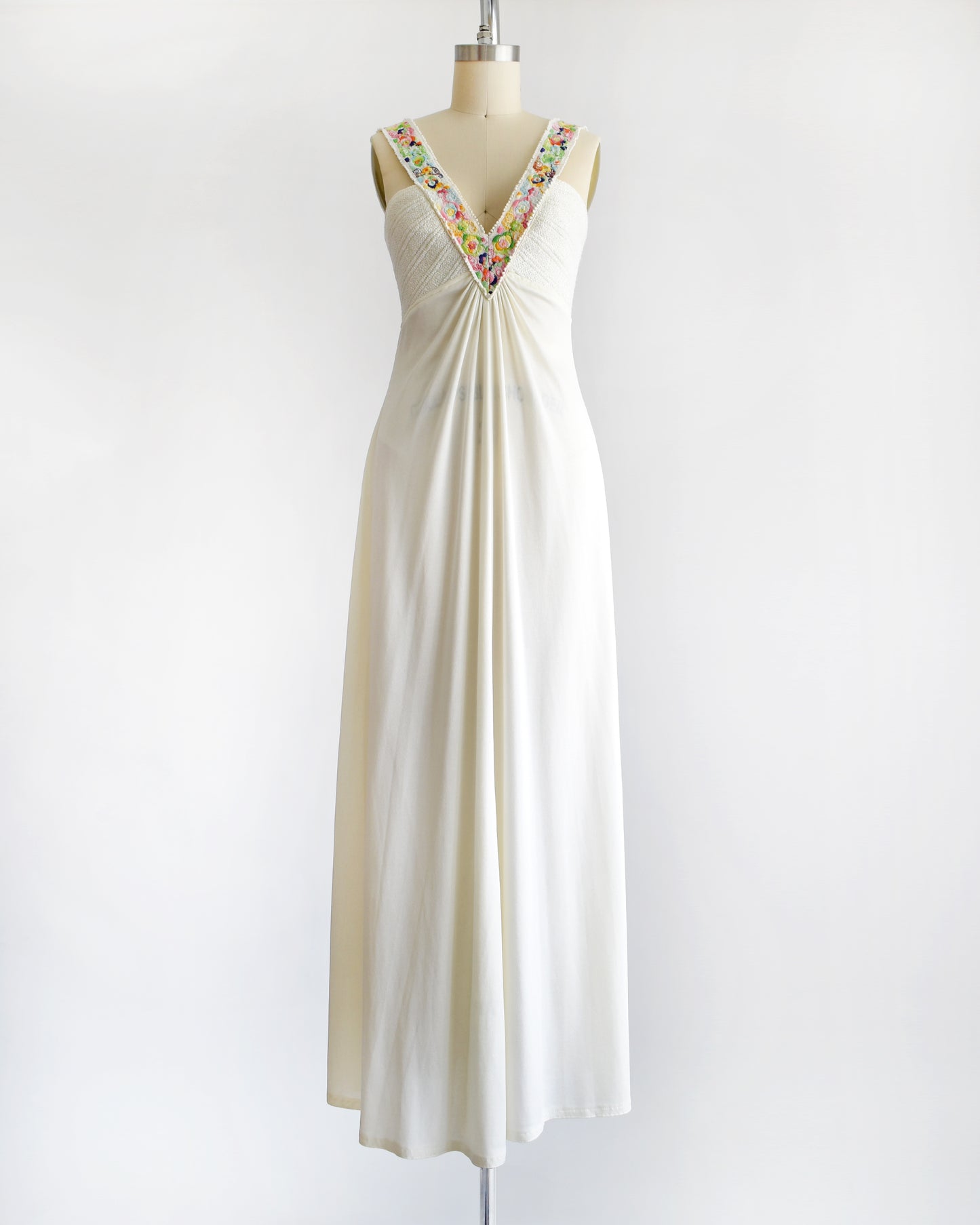 a vintage 1970s white maxi dress with colorful floral embroidery on the straps that go down the front on a dress form against a white background