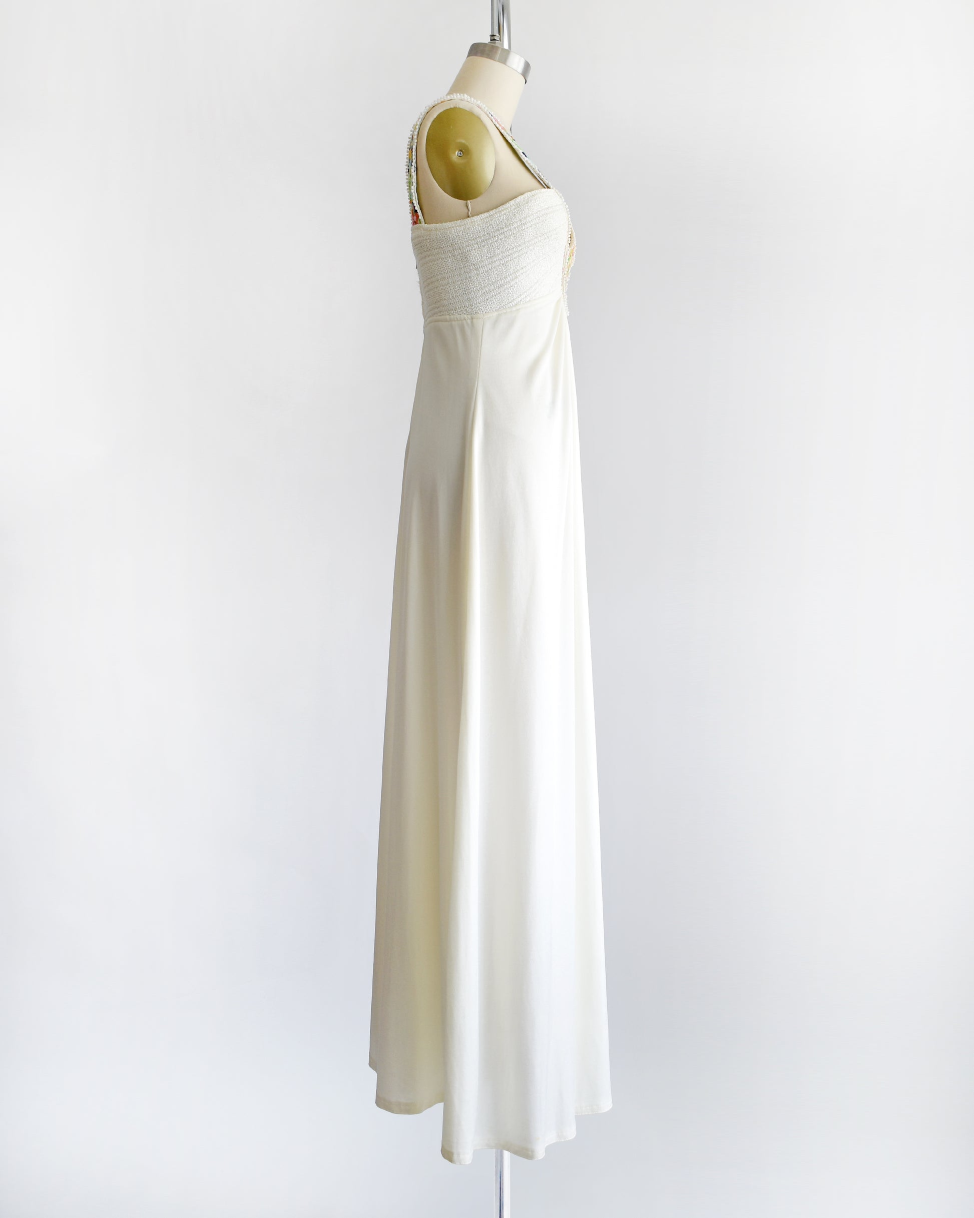 side view of a vintage 1970s white maxi dress with colorful floral embroidery on the straps that go down the front on a dress form against a white background