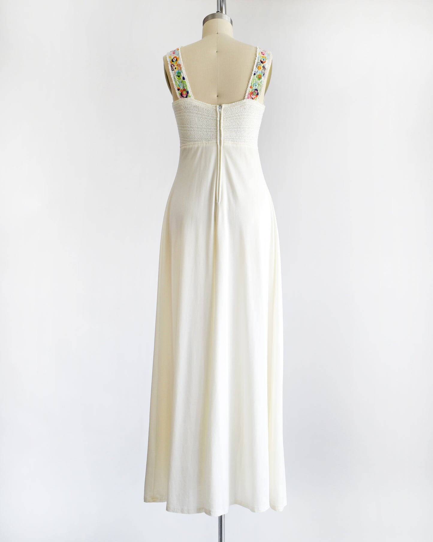 a vintage 1970s white maxi dress with colorful floral embroidery on the straps on a dress form against a white background