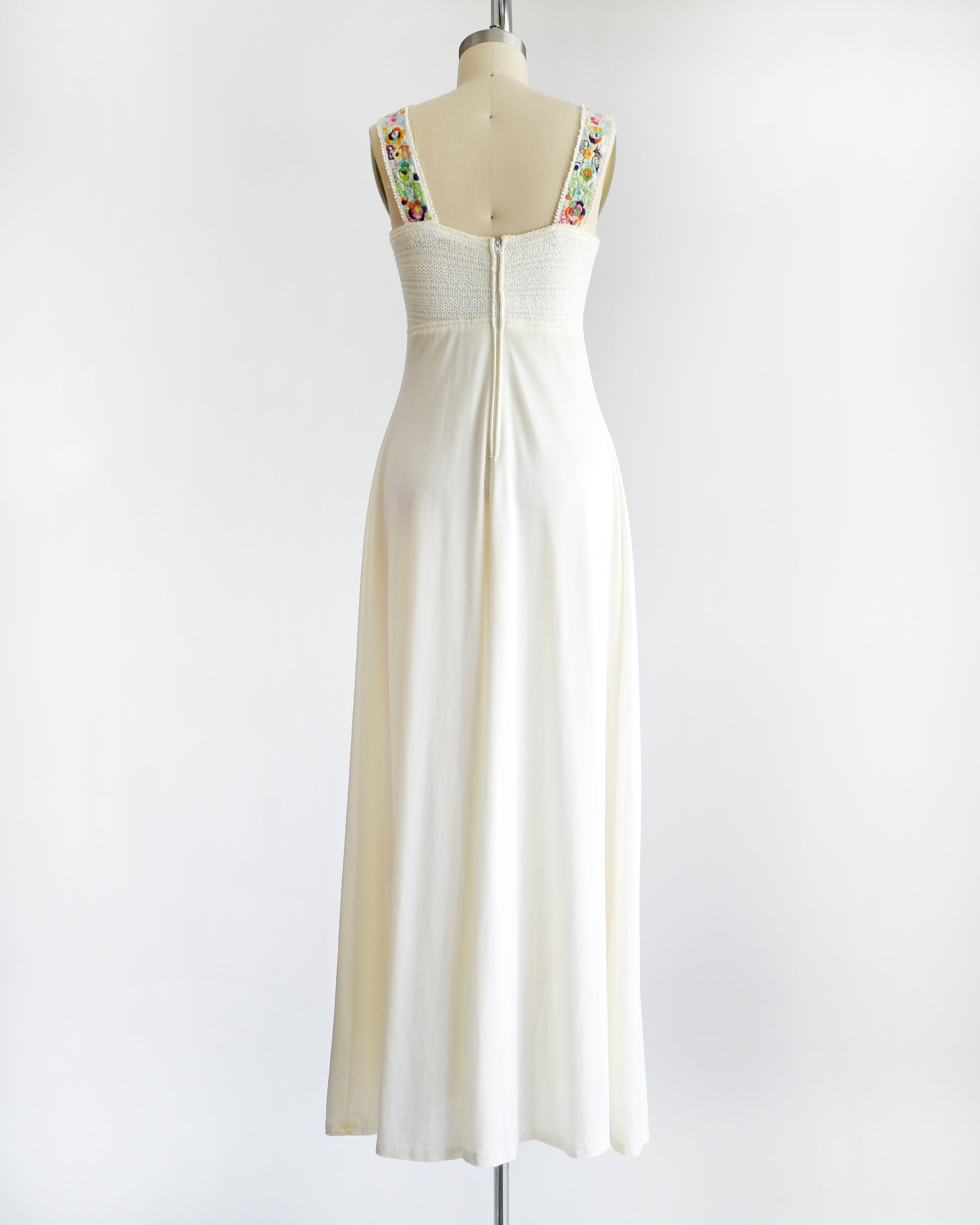 a vintage 1970s white maxi dress with colorful floral embroidery on the straps on a dress form against a white background