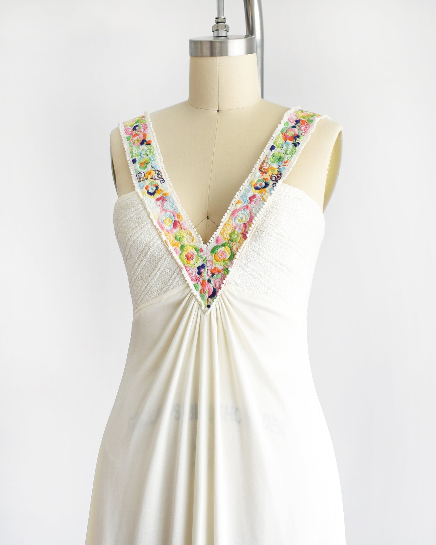 side front view of a vintage 1970s white maxi dress with colorful floral embroidery on the straps that go down the front on a dress form against a white background