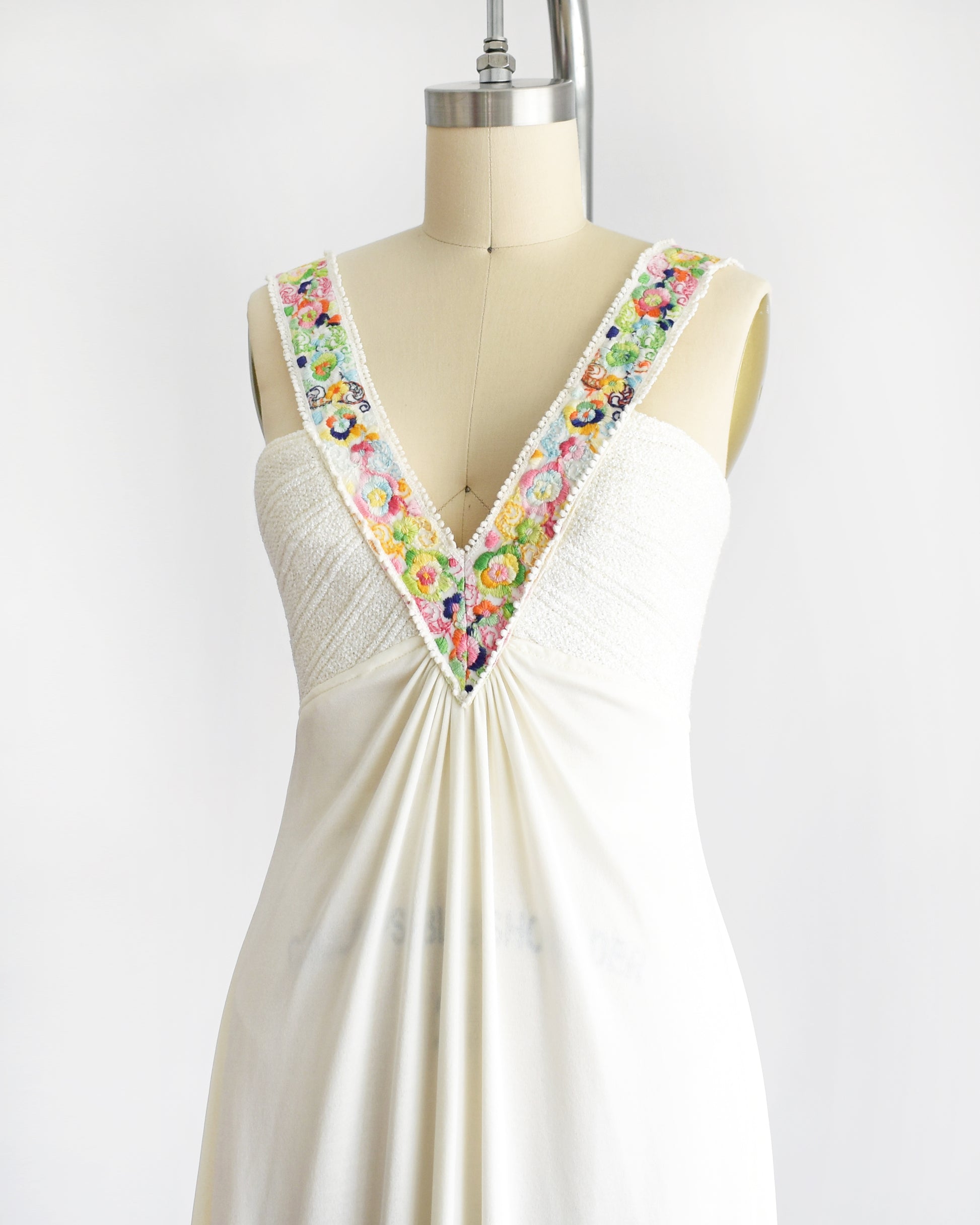side front view of a vintage 1970s white maxi dress with colorful floral embroidery on the straps that go down the front on a dress form against a white background