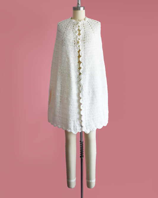 a vintage 1970s white poncho with scalloped trim on a mannequin against a pink background