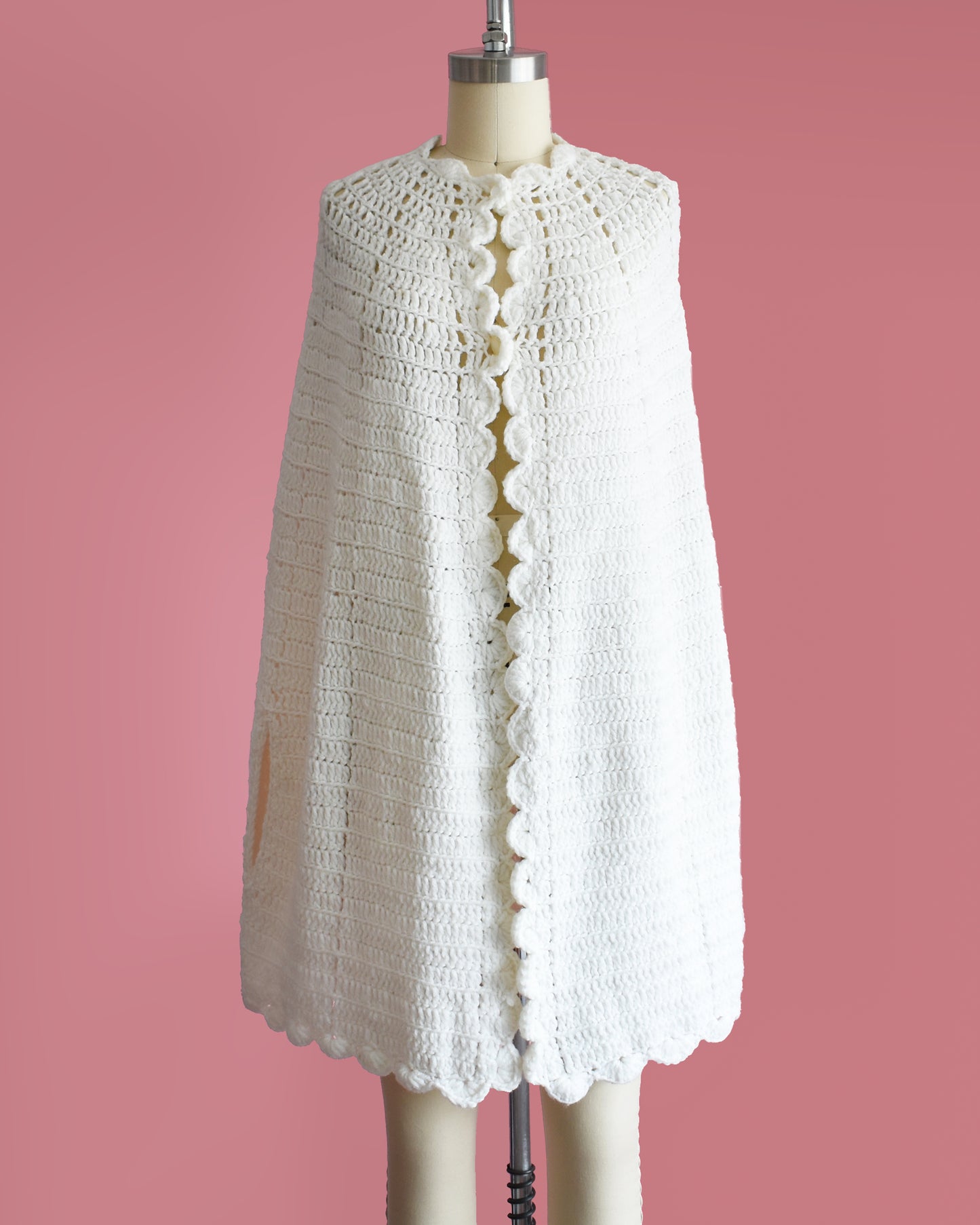 a vintage 1970s white poncho with scalloped trim on a mannequin against a pink background