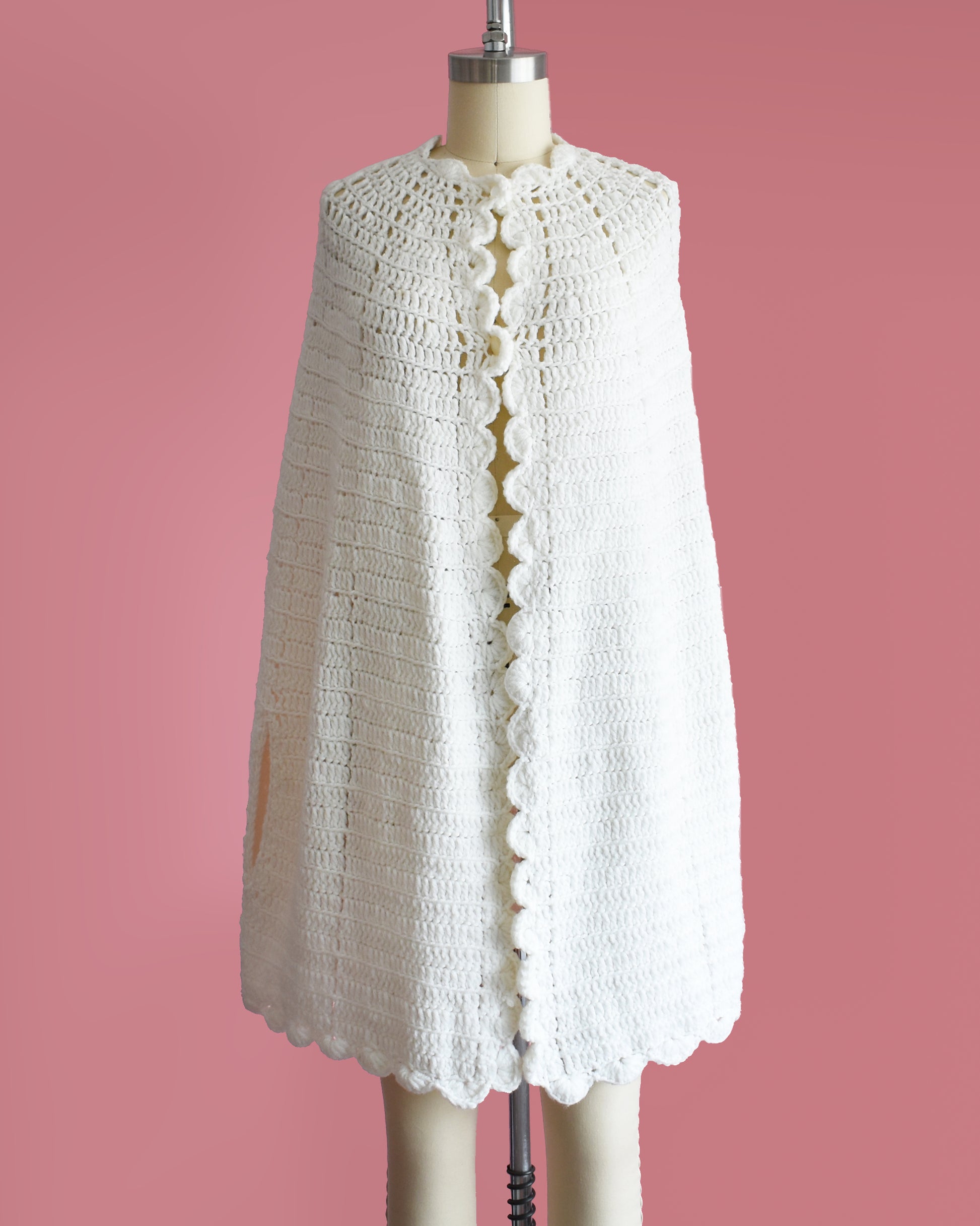 a vintage 1970s white poncho with scalloped trim on a mannequin against a pink background