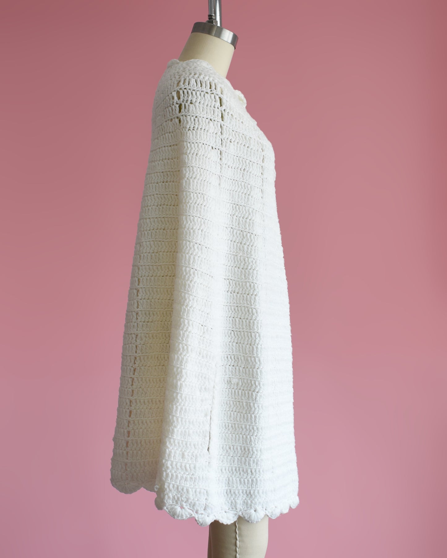 side view of a vintage 1970s white poncho with scalloped trim on a mannequin against a pink background
