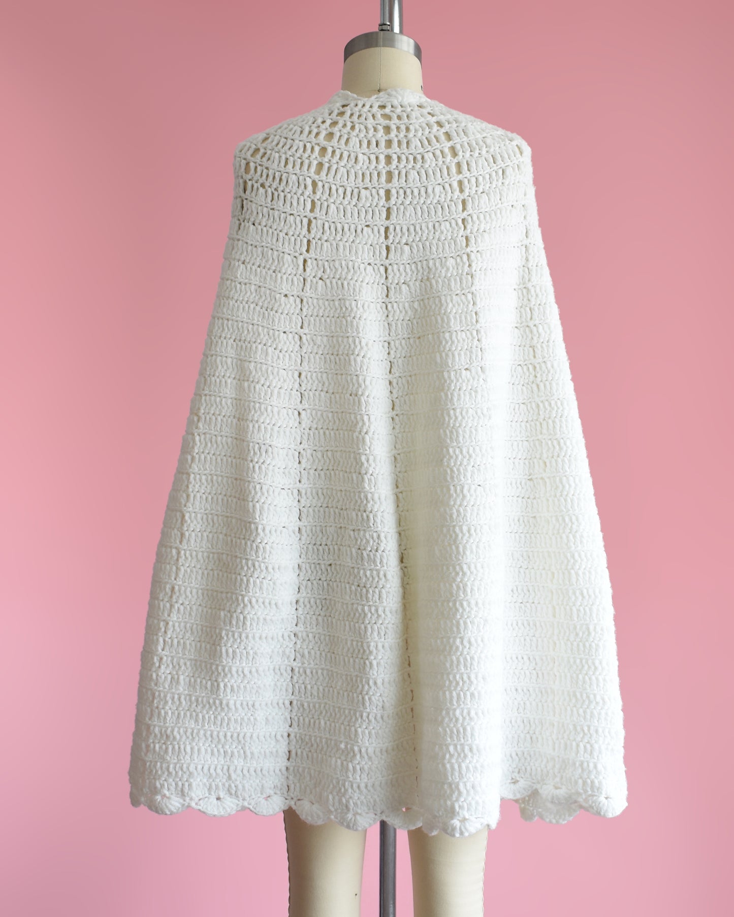back view of a vintage 1970s white poncho with scalloped trim on a mannequin against a pink background