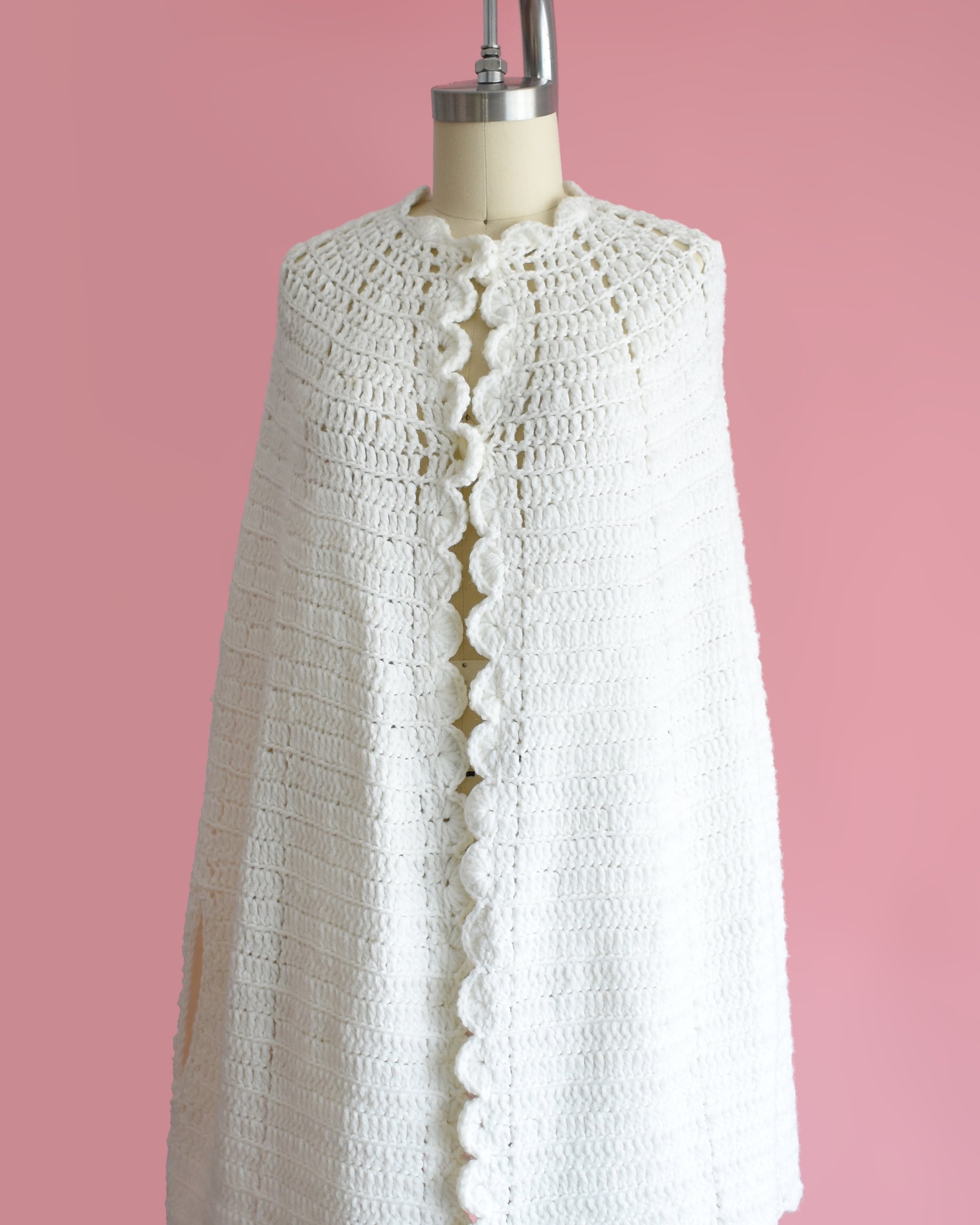 side front view of a vintage 1970s white poncho with scalloped trim on a mannequin against a pink background