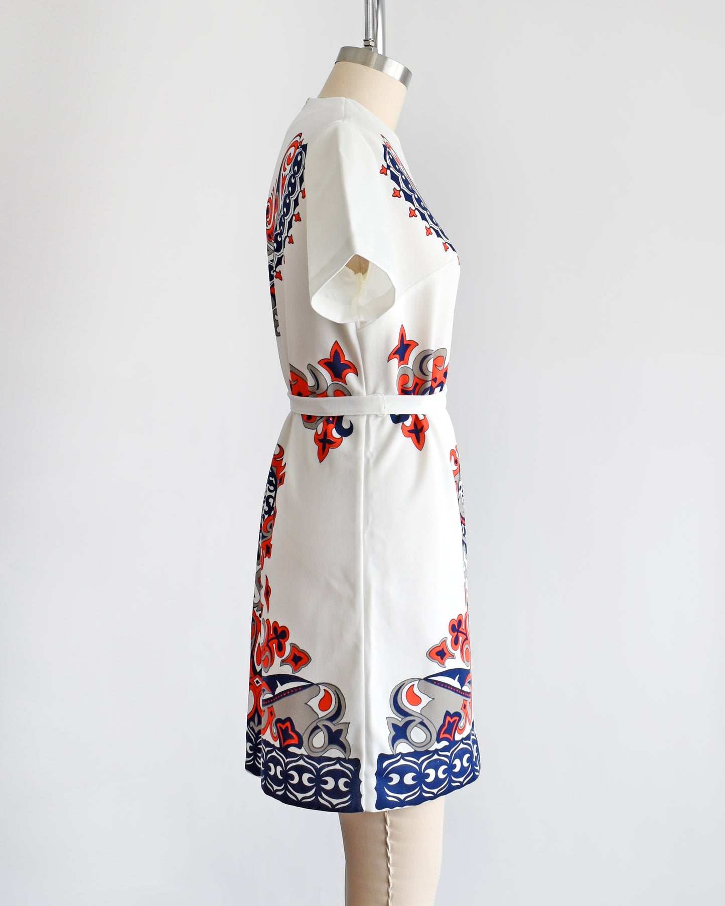 side view of a vintage 1970s white mod dress that has a red, navy blue, and gray damask print on the front, sides, and back. the dress comes with a matching white tie belt