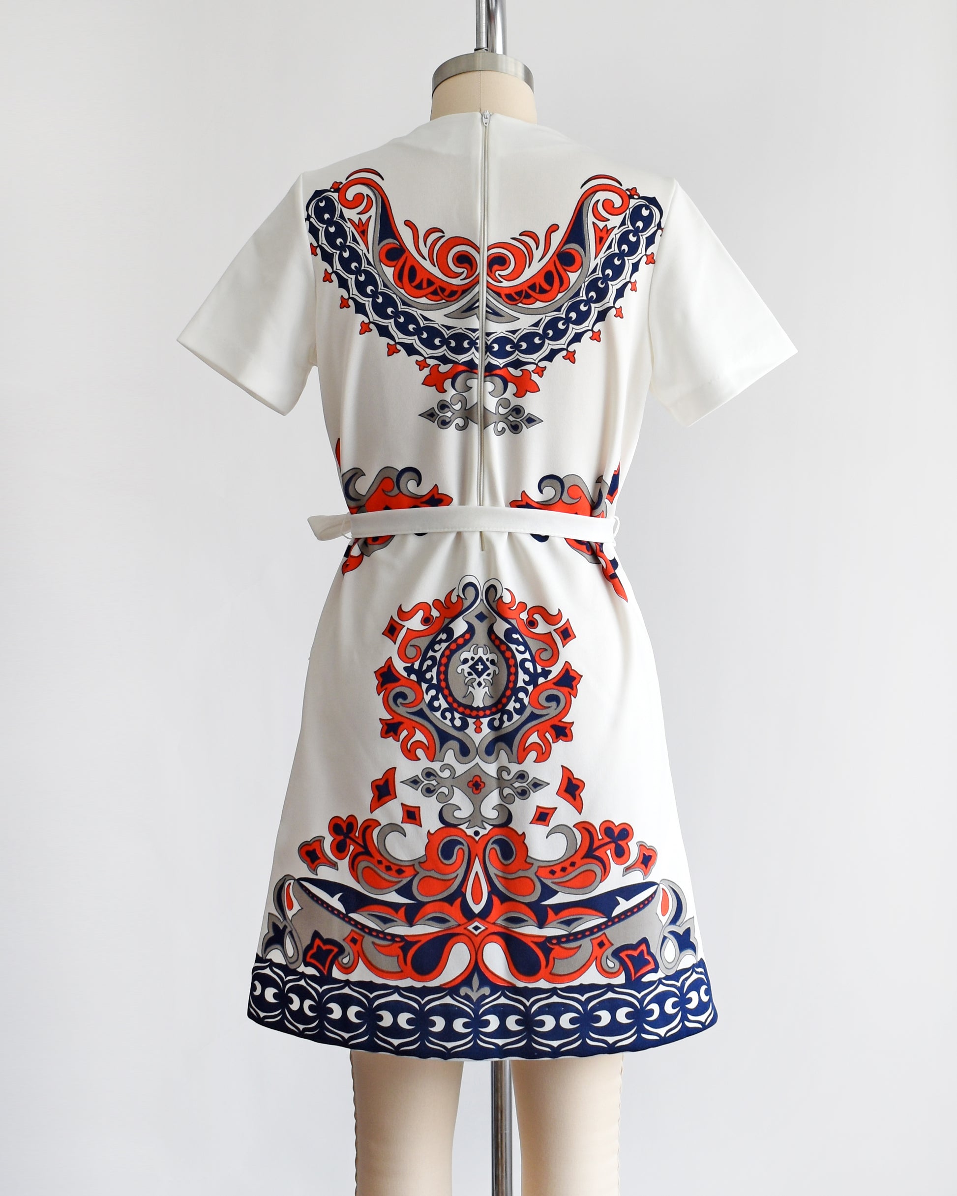 back view of a vintage 1970s white mod dress that has a red, navy blue, and gray damask print on the front, sides, and back. the dress comes with a matching white tie belt