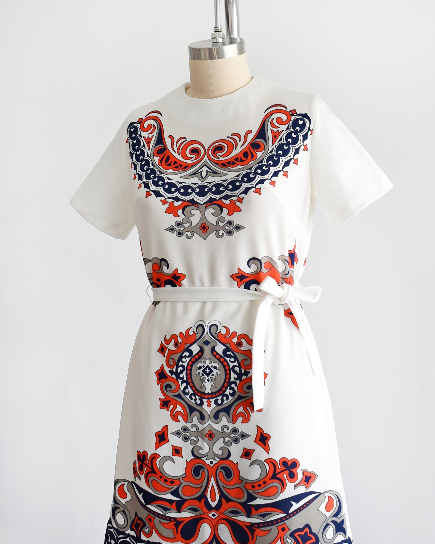 side front view of a vintage 1970s white mod dress that has a red, navy blue, and gray damask print on the front, sides, and back. the dress comes with a matching white tie belt