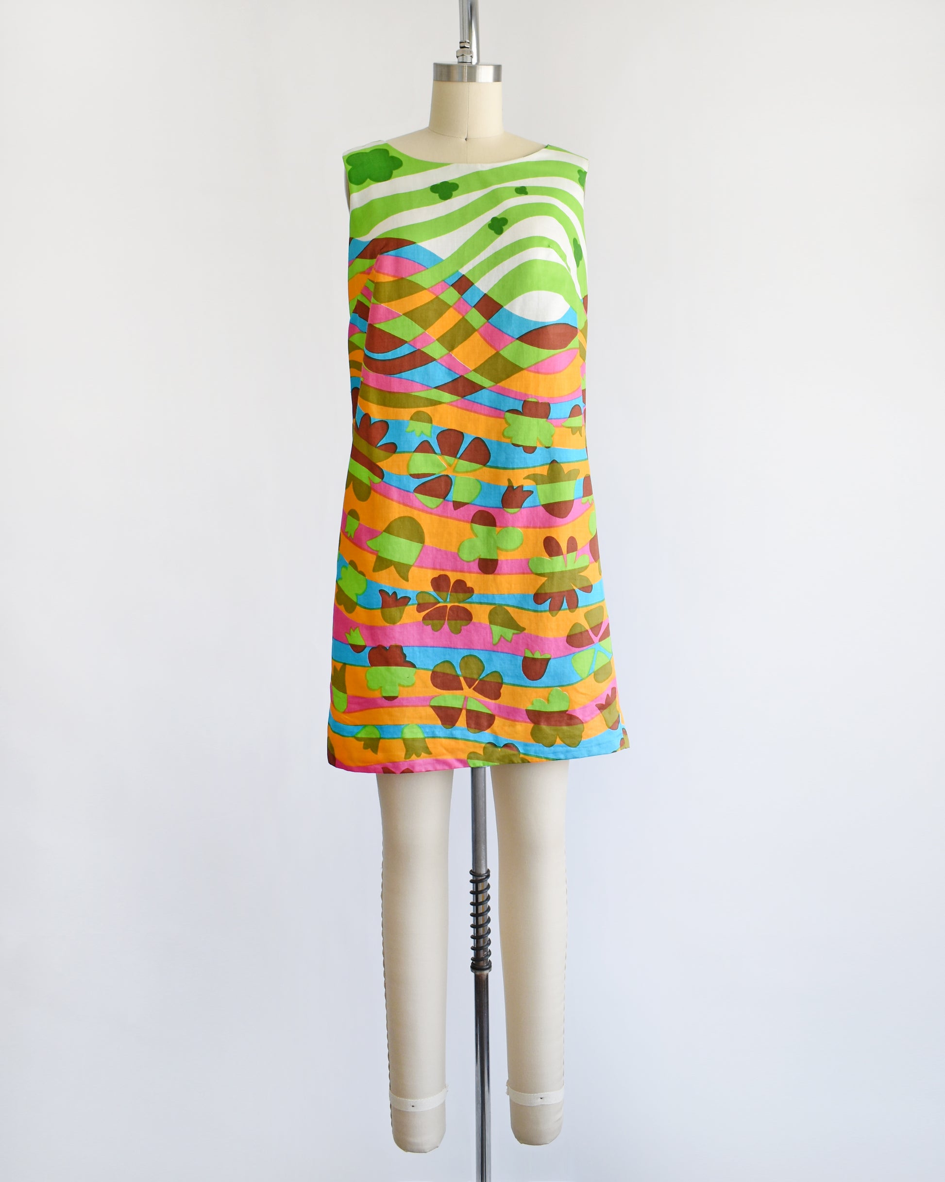 A vintage 1960s sleeveless dress displayed on a cream mannequin against a plain white background. The dress features a vibrant, multicolored abstract pattern with wavy horizontal bands in green, pink, orange, blue, yellow, and brown.