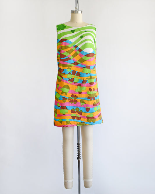 A vintage 1960s sleeveless dress displayed on a cream mannequin against a plain white background. The dress features a vibrant, multicolored abstract pattern with wavy horizontal bands in green, pink, orange, blue, yellow, and brown.