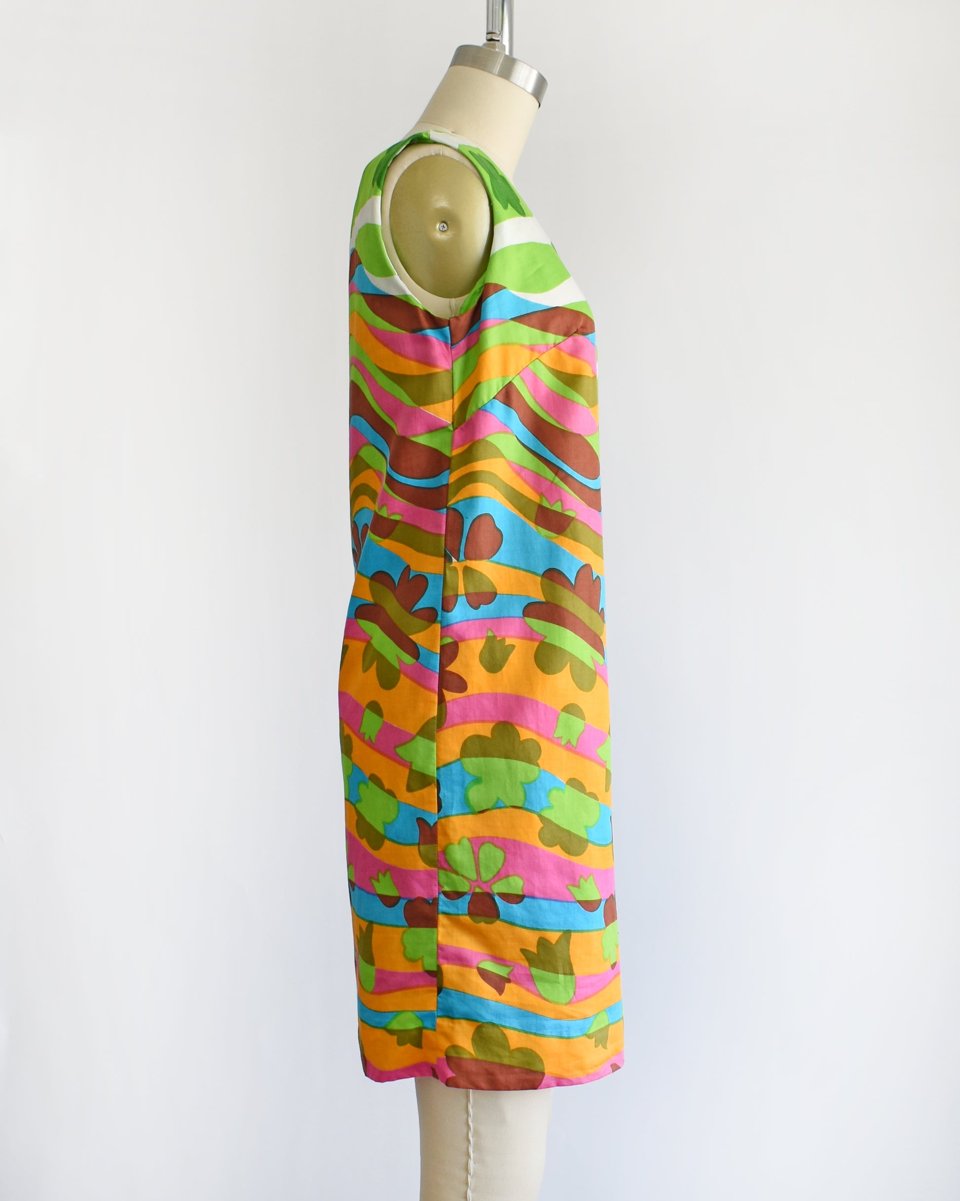 side view of a vintage 1960s sleeveless dress displayed on a cream mannequin against a plain white background. The dress features a vibrant, multicolored abstract pattern with wavy horizontal bands in green, pink, orange, blue, yellow, and brown.