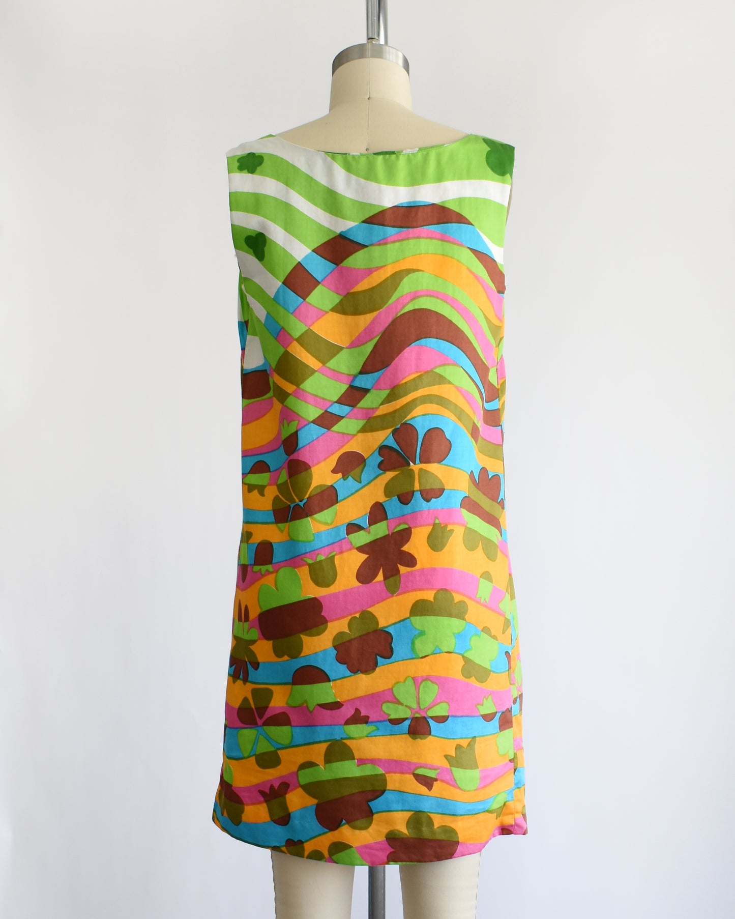 back view of a vintage 1960s sleeveless dress displayed on a cream mannequin against a plain white background. The dress features a vibrant, multicolored abstract pattern with wavy horizontal bands in green, pink, orange, blue, yellow, and brown.