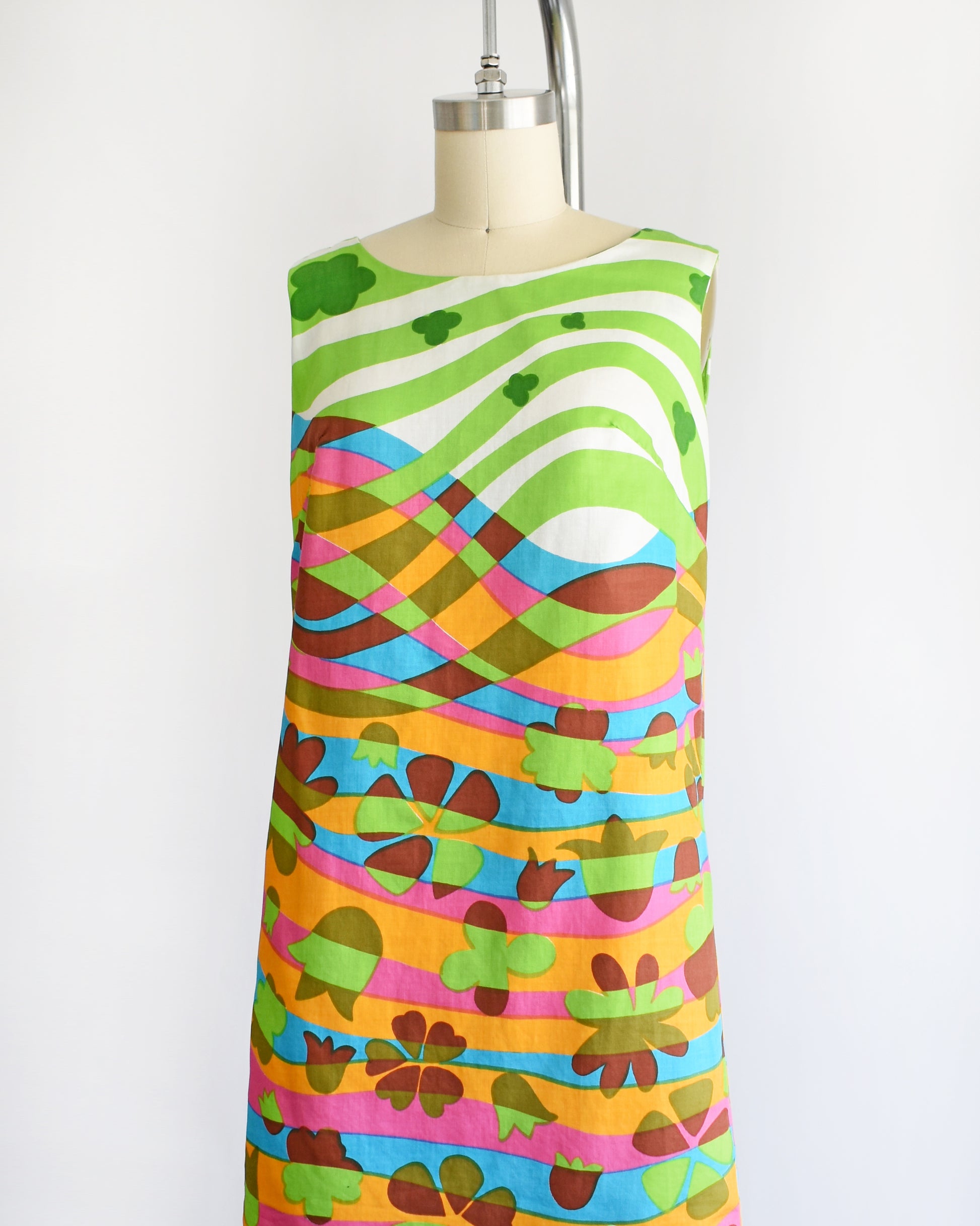 side front view of a vintage 1960s sleeveless dress displayed on a cream mannequin against a plain white background. The dress features a vibrant, multicolored abstract pattern with wavy horizontal bands in green, pink, orange, blue, yellow, and brown.