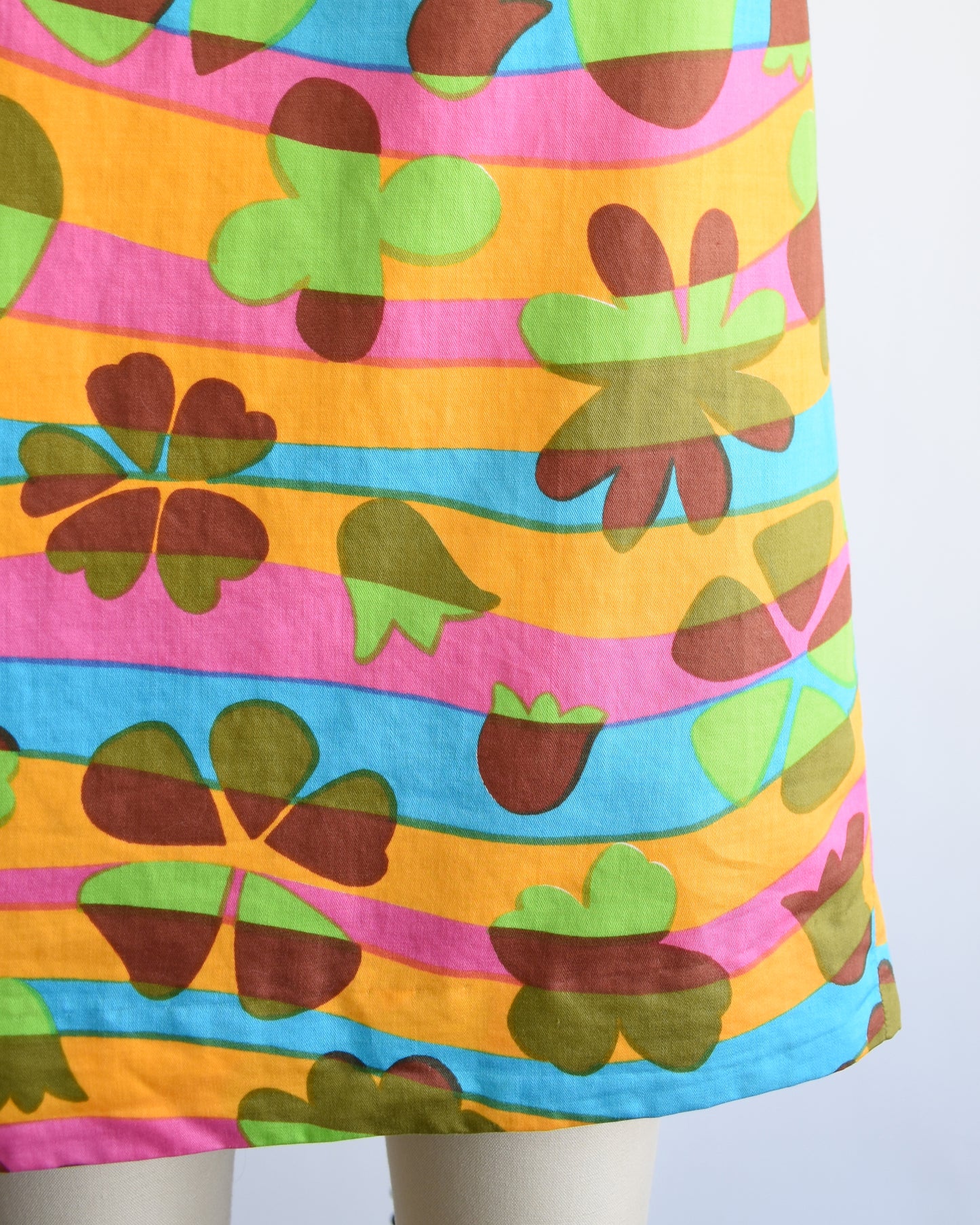 close up of the hem of a vintage 1960s sleeveless dress displayed on a cream mannequin against a plain white background. The dress features a vibrant, multicolored abstract pattern with wavy horizontal bands in green, pink, orange, blue, yellow, and brown.