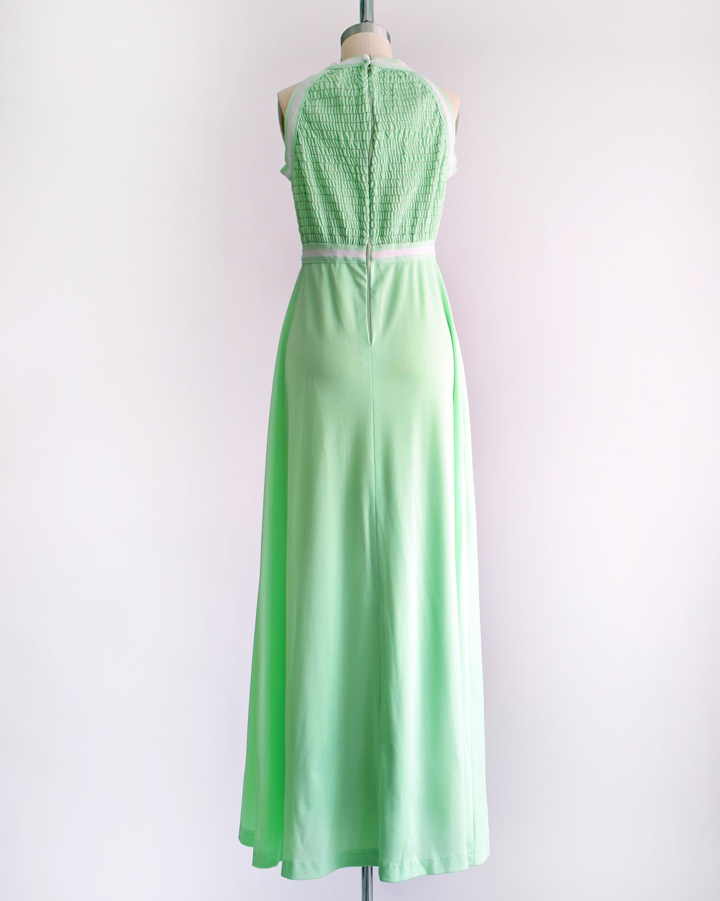 back view of a vintage 1970s mint green sleeveless halter maxi dress with a smocked bodice and a flared skirt, displayed on a dress form against a plain light background.